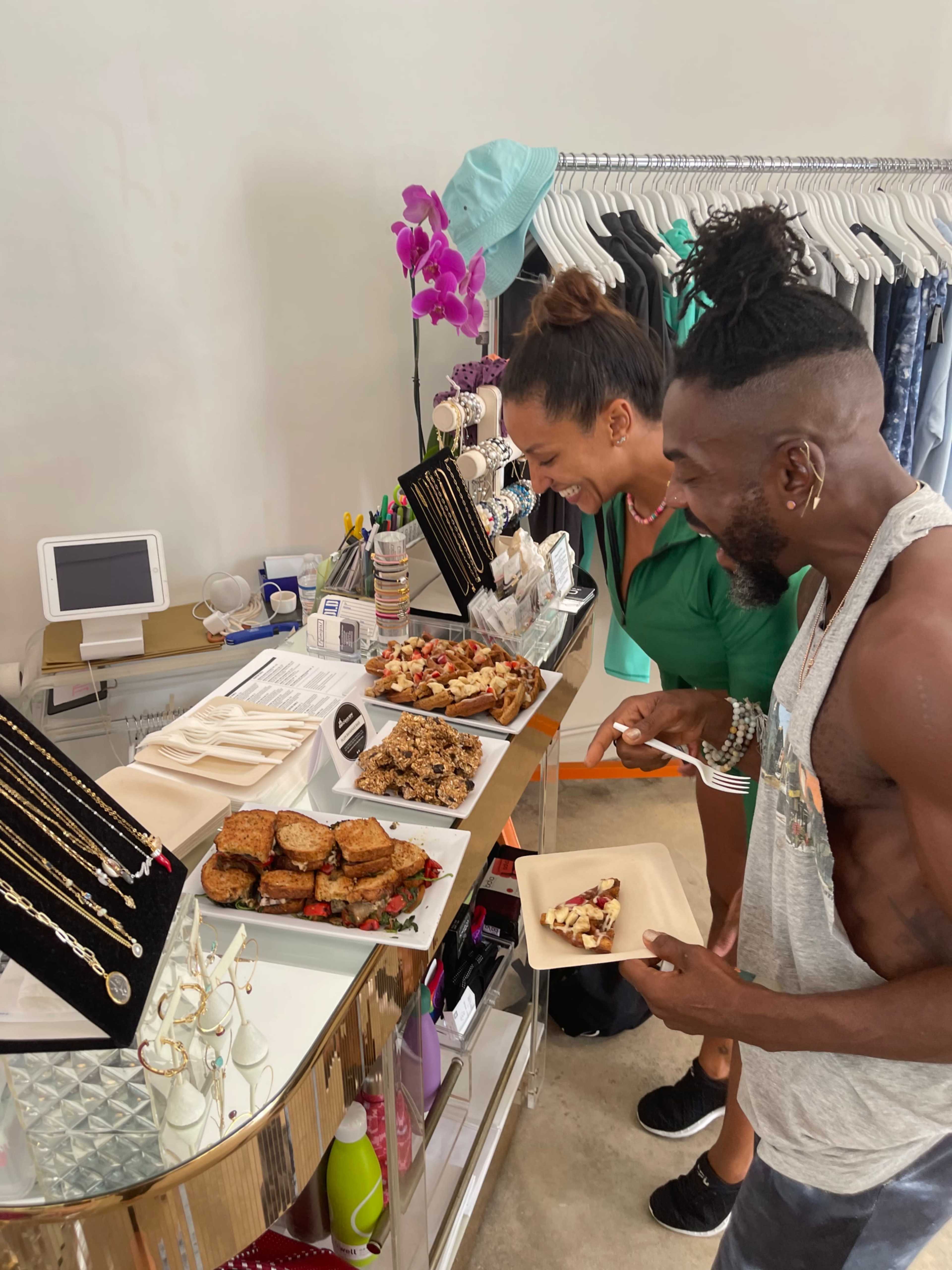 Two people are sampling various food items displayed on a table in a boutique setting.
