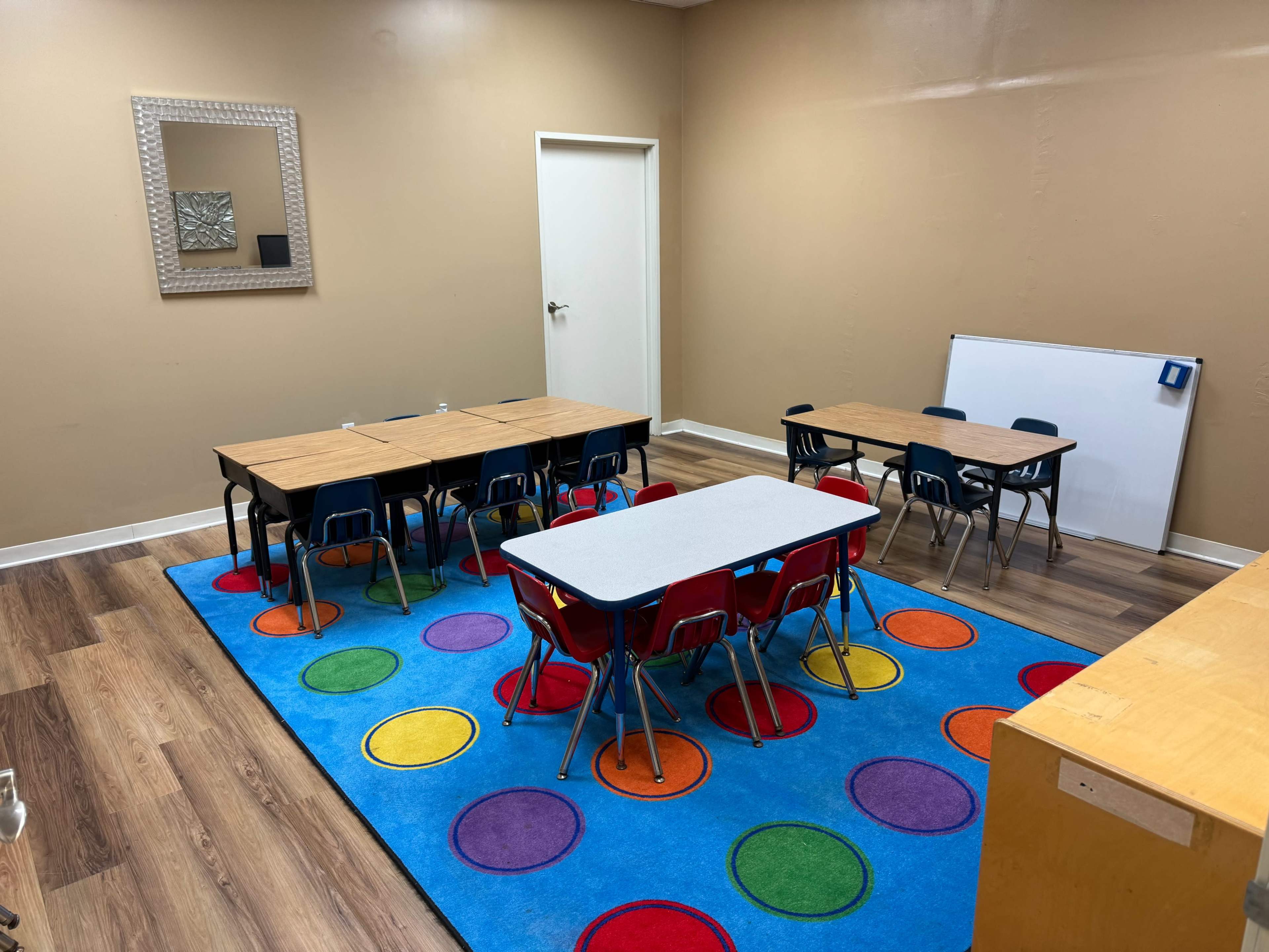 A classroom with multiple wooden tables and chairs arranged on a colorful carpet, along with a whiteboard and a mirror on the wall.