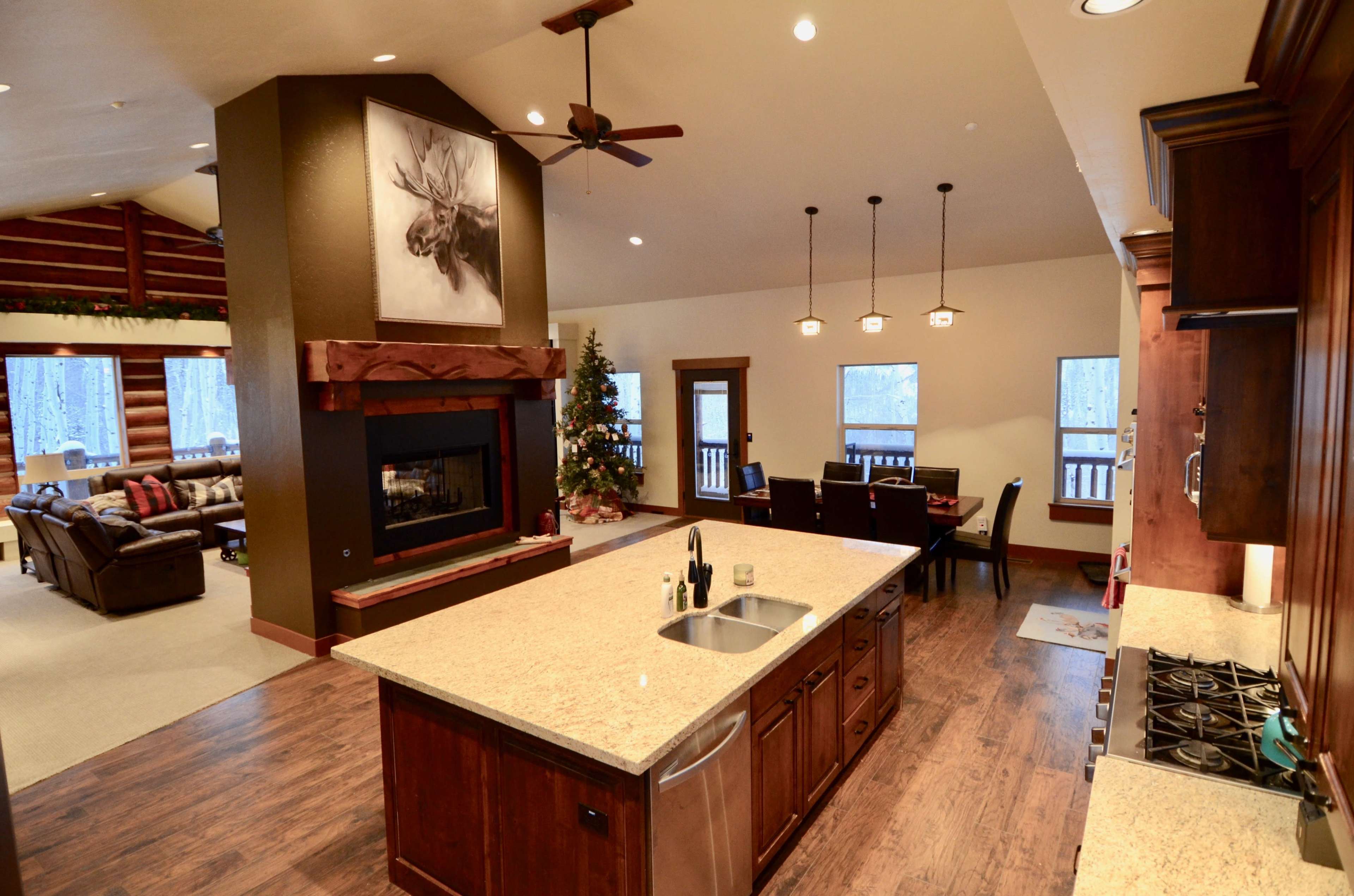 An open-concept living space featuring a kitchen island with a sink, a dining area with a wooden table, and a cozy living room with a fireplace and holiday decor.