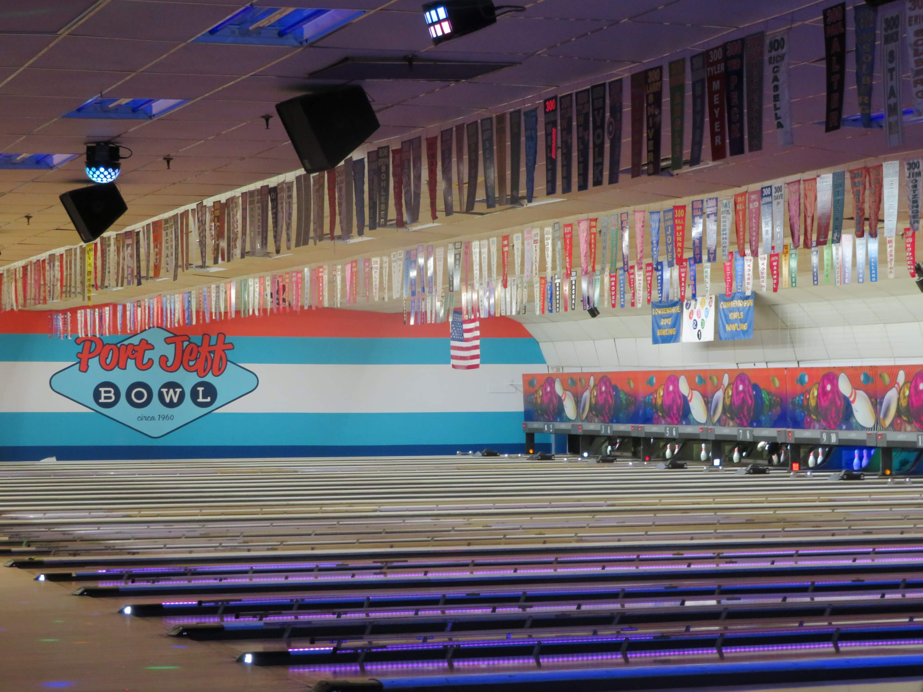 The image shows the interior of Port Jeff Bowl, with numerous bowling lanes and banners hanging from the ceiling.