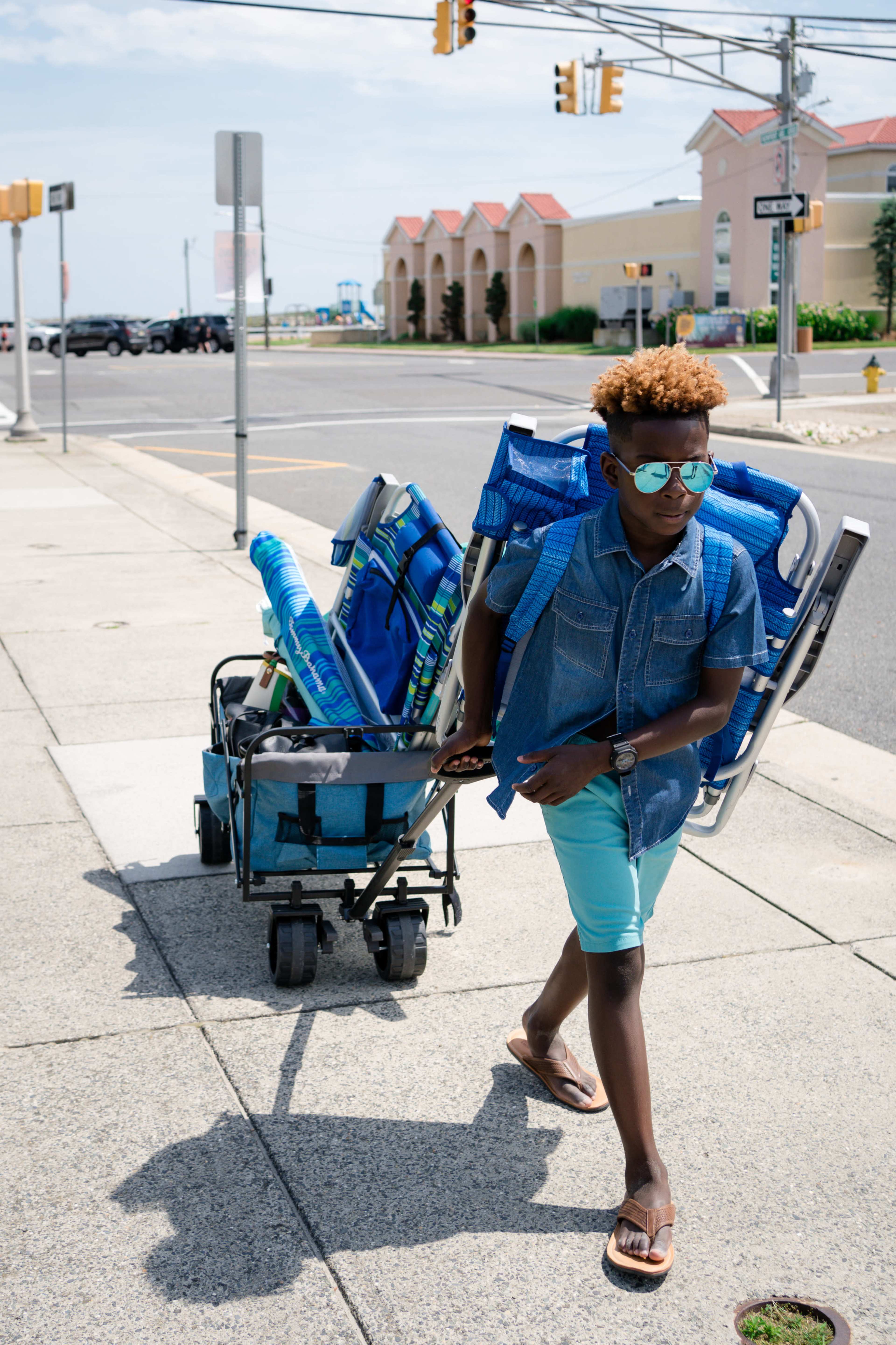 A child walks down the sidewalk, pulling a cart filled with beach chairs and wearing sunglasses.