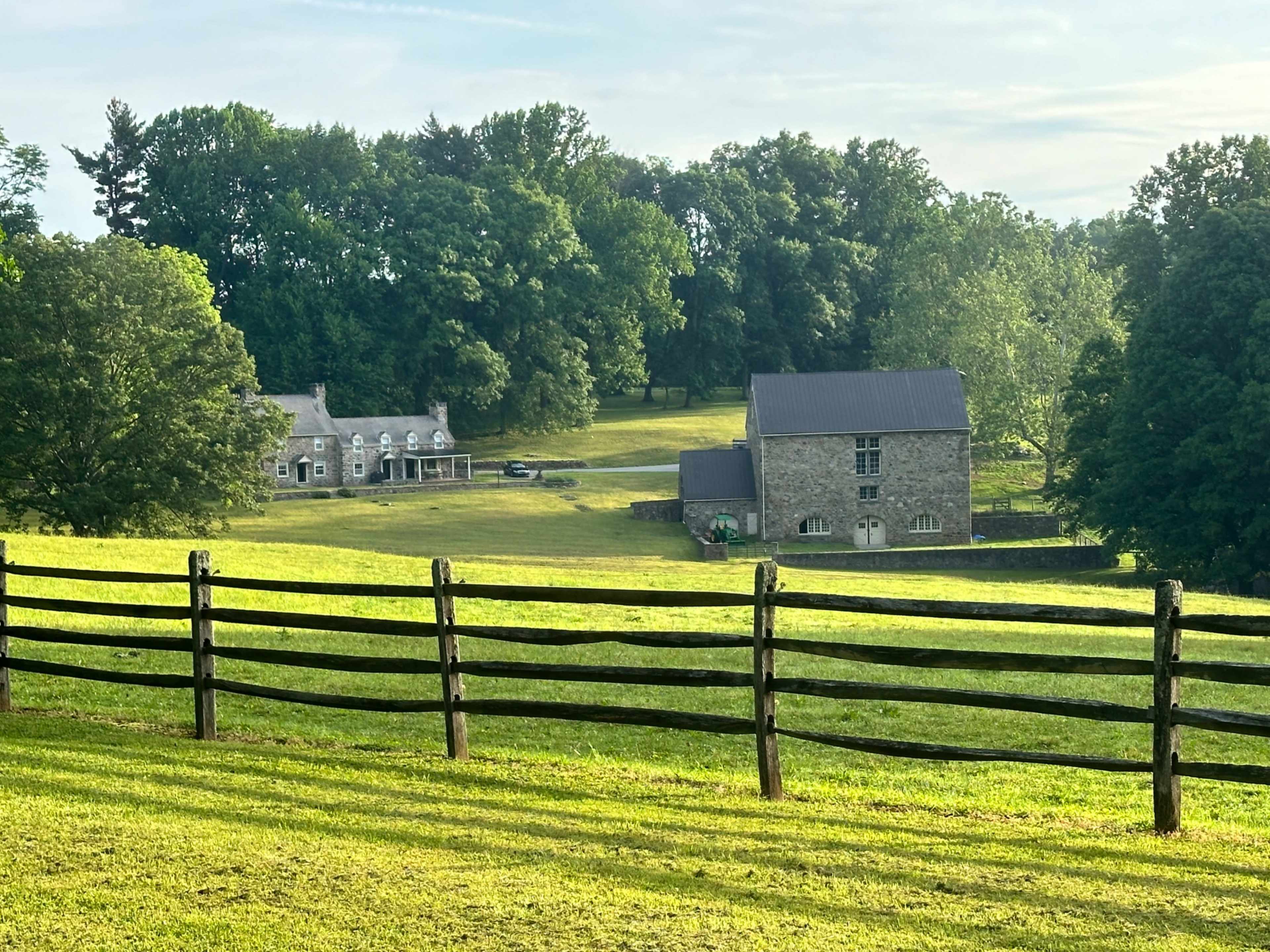 The image shows a stone building and a house set against a backdrop of green trees and a wooden fence in a rural landscape.
