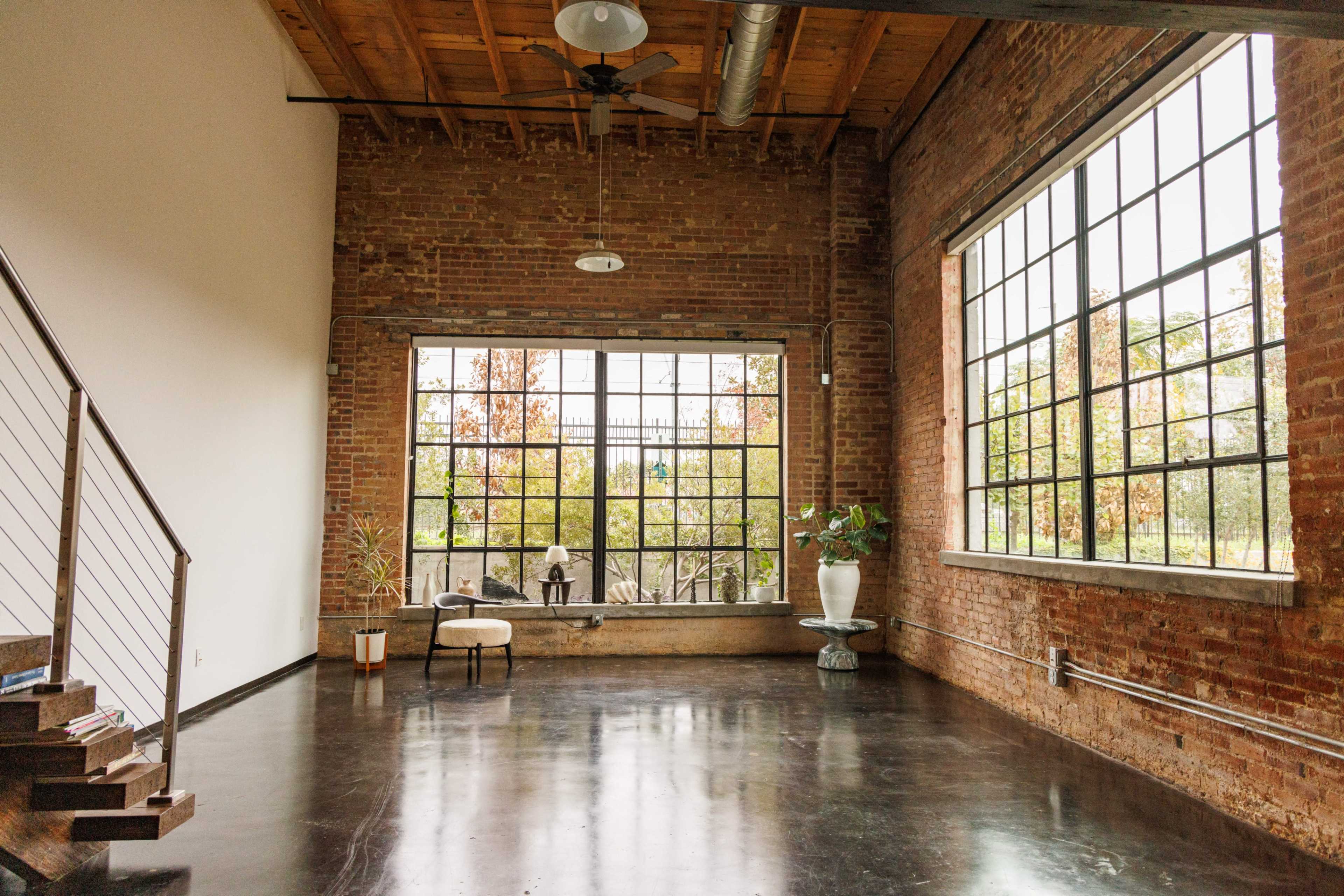 The image shows an empty industrial-style room with exposed brick walls, large windows, and a staircase in the corner.