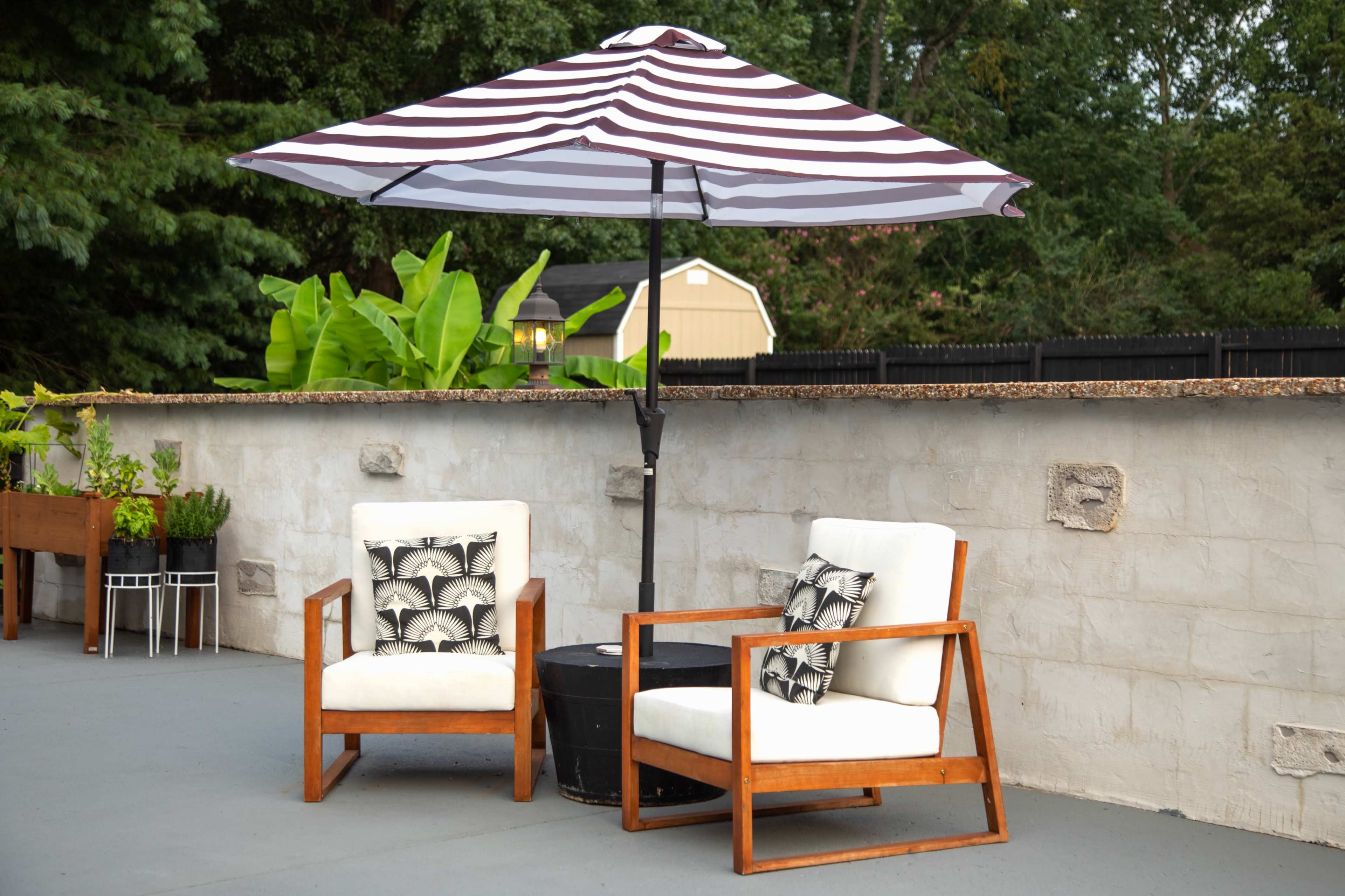 Two wooden chairs with white cushions and patterned pillows positioned under a large striped umbrella on a patio, adjacent to a stone wall and greenery.