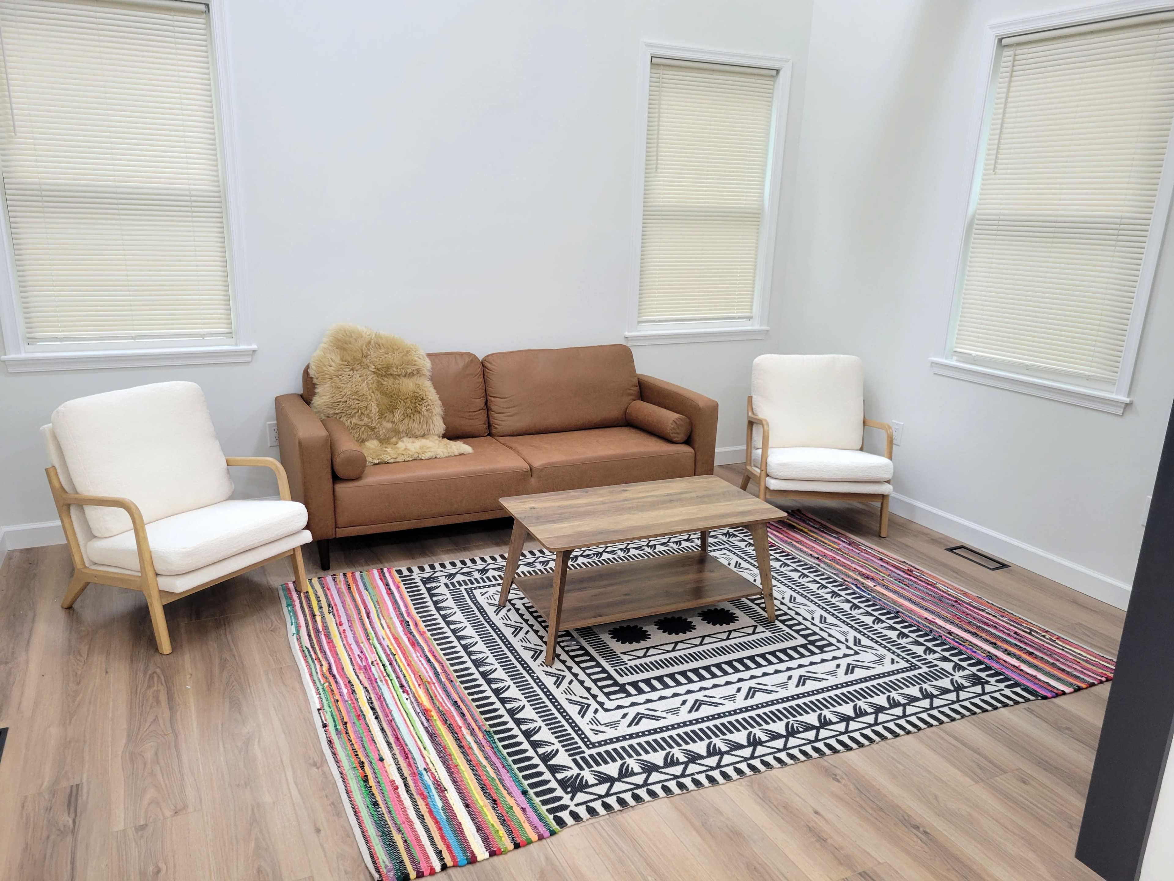 The image shows a light, modern living room with a brown sofa, two white armchairs, a wooden coffee table, and a colorful area rug featuring geometric patterns.