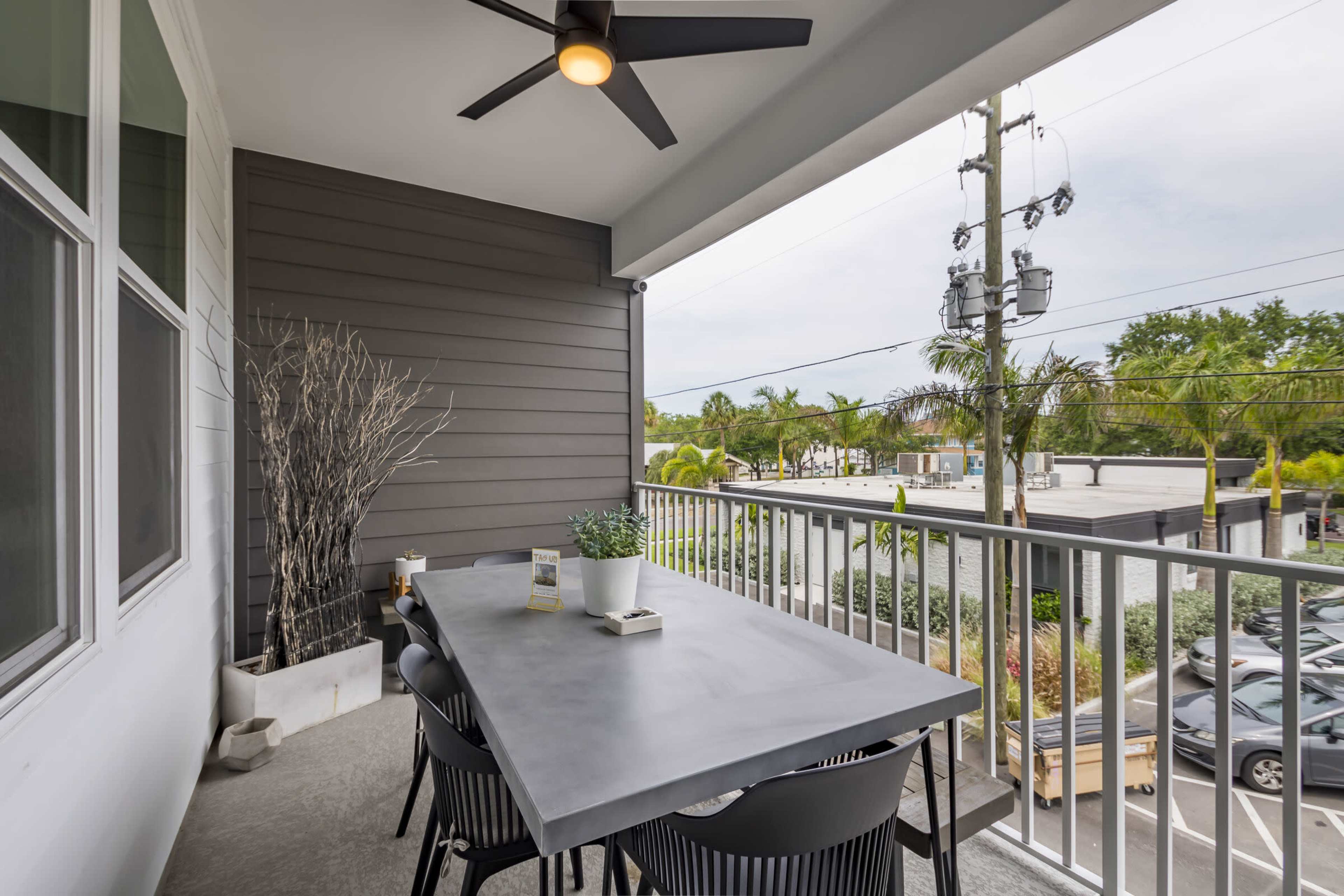 The image shows a spacious balcony with a concrete dining table and black chairs, surrounded by greenery and power lines.