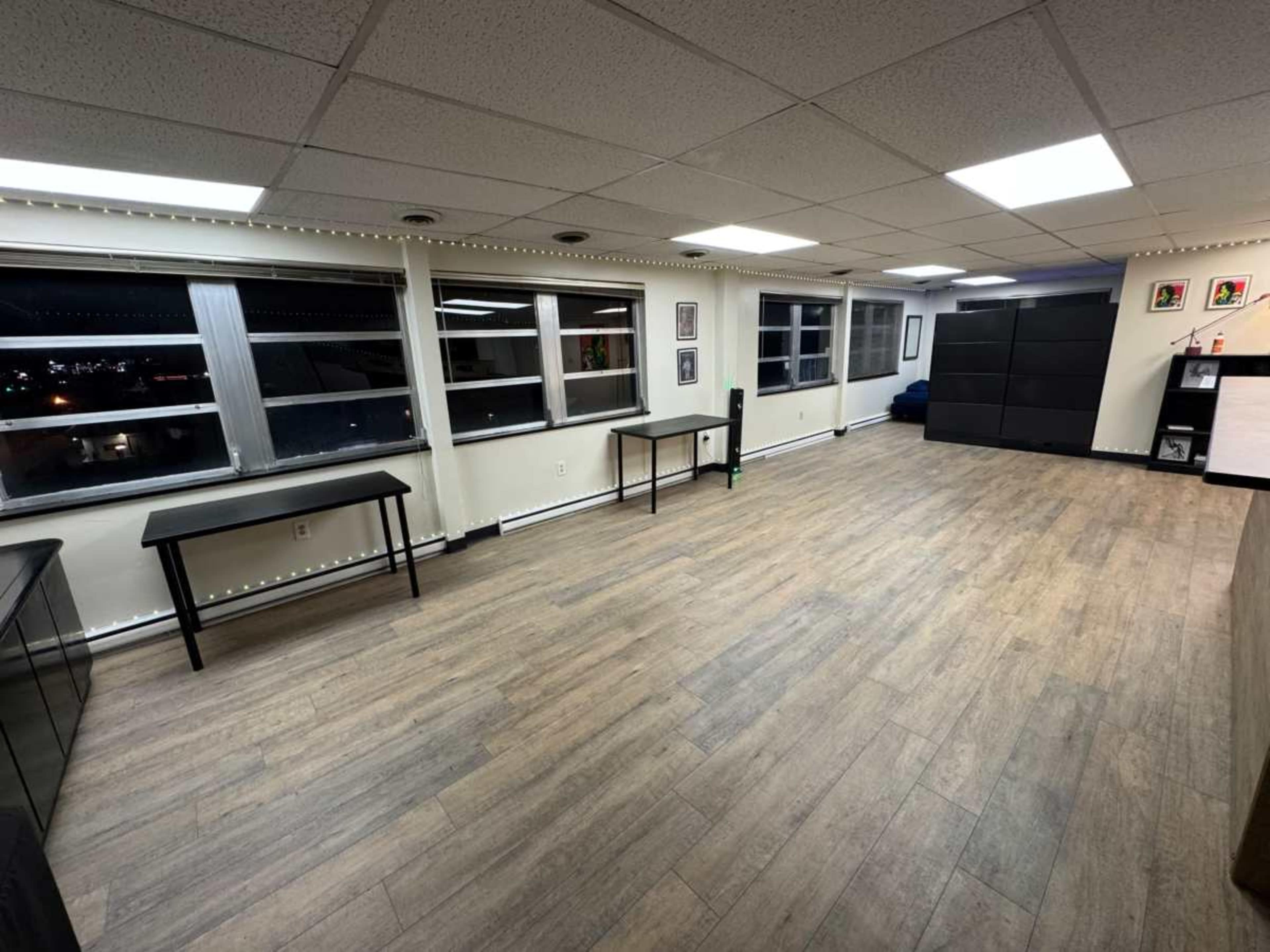 The image shows a spacious, well-lit room with large windows, a light brown wood floor, and minimal furniture, including a couple of tables and storage units.