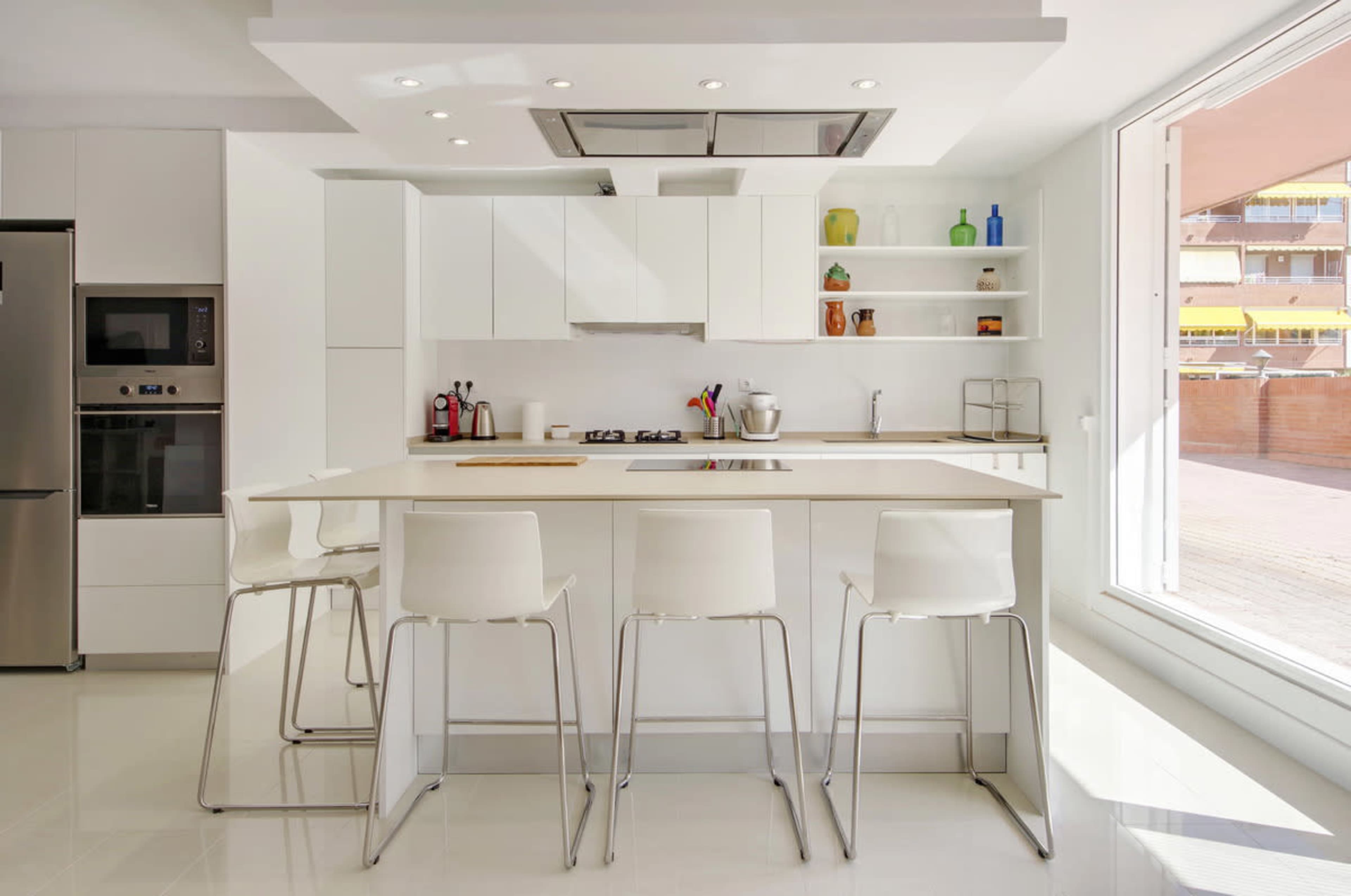 A modern kitchen features a central island with four white bar stools, light countertops, and sleek cabinetry against a bright, airy background.