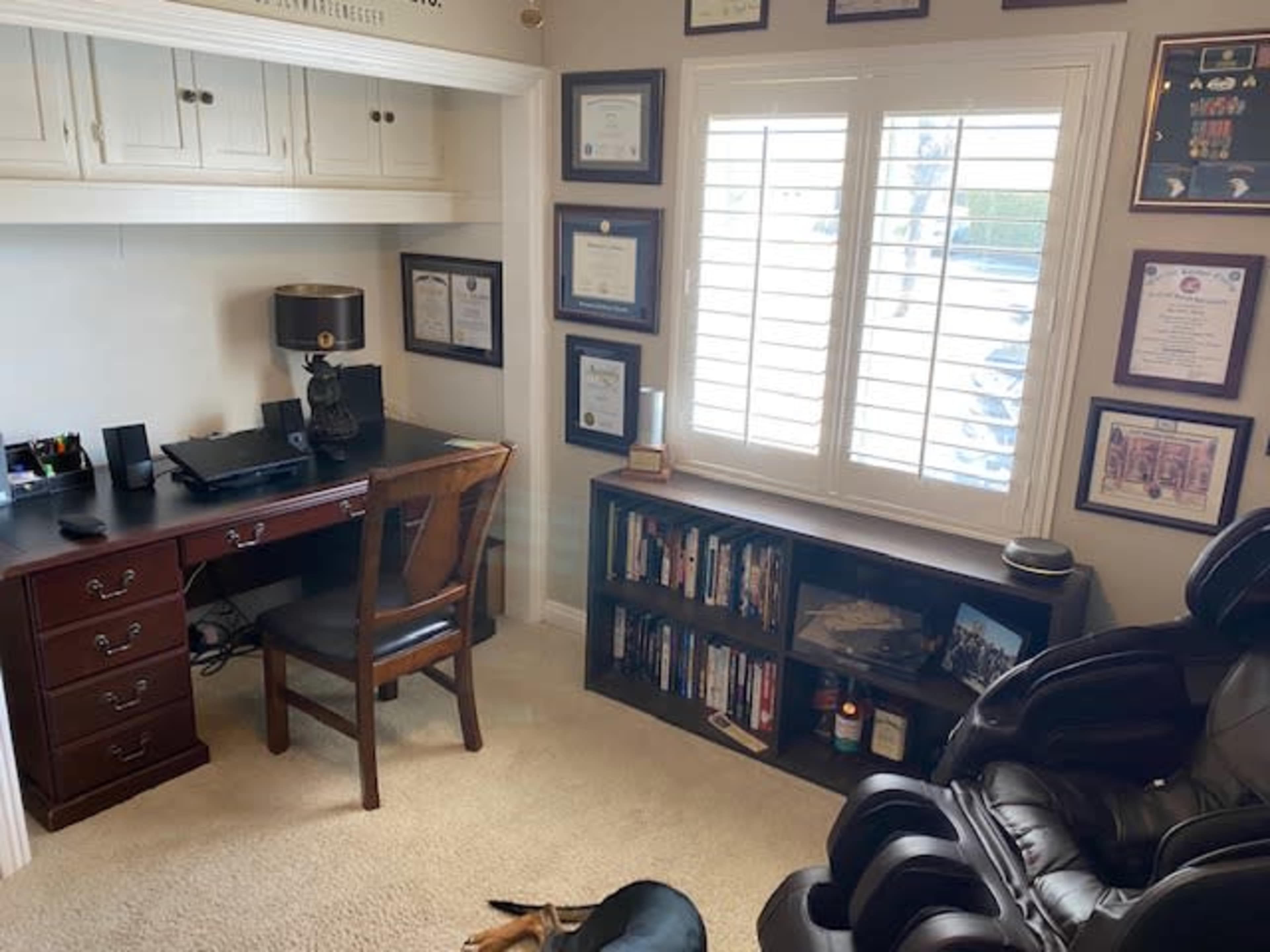 A home office with a wooden desk, a chair, a mounted lamp, and shelves filled with books and framed certificates on the walls.