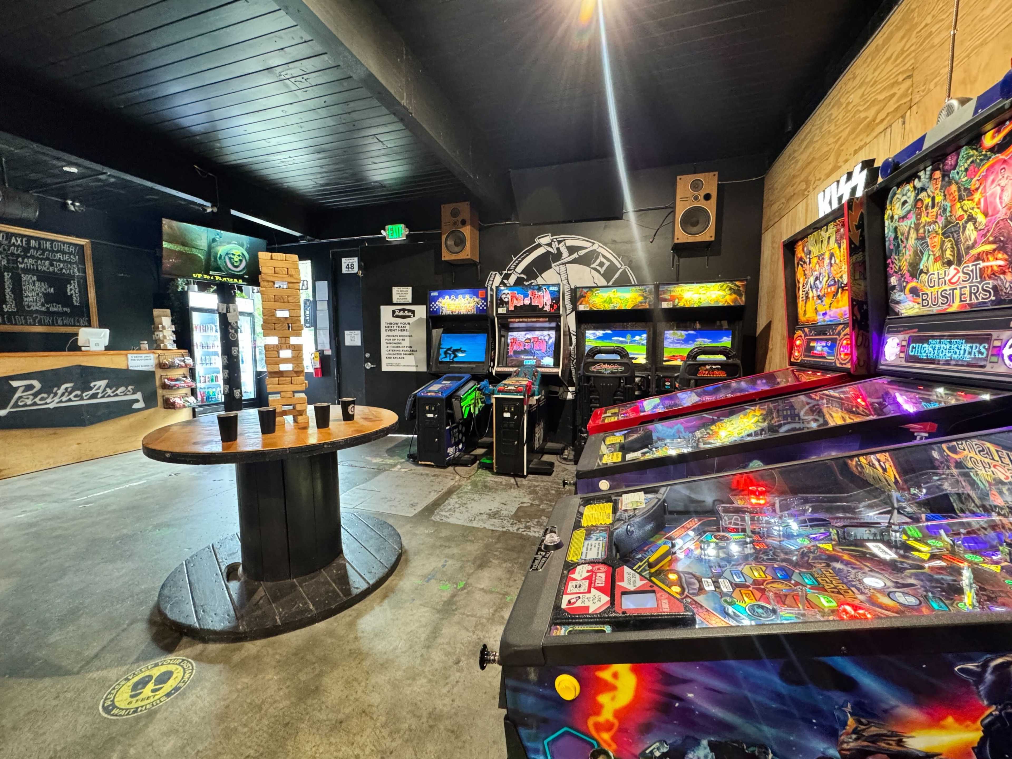 The image shows a well-lit arcade featuring several pinball machines and classic arcade games arranged around a central table.
