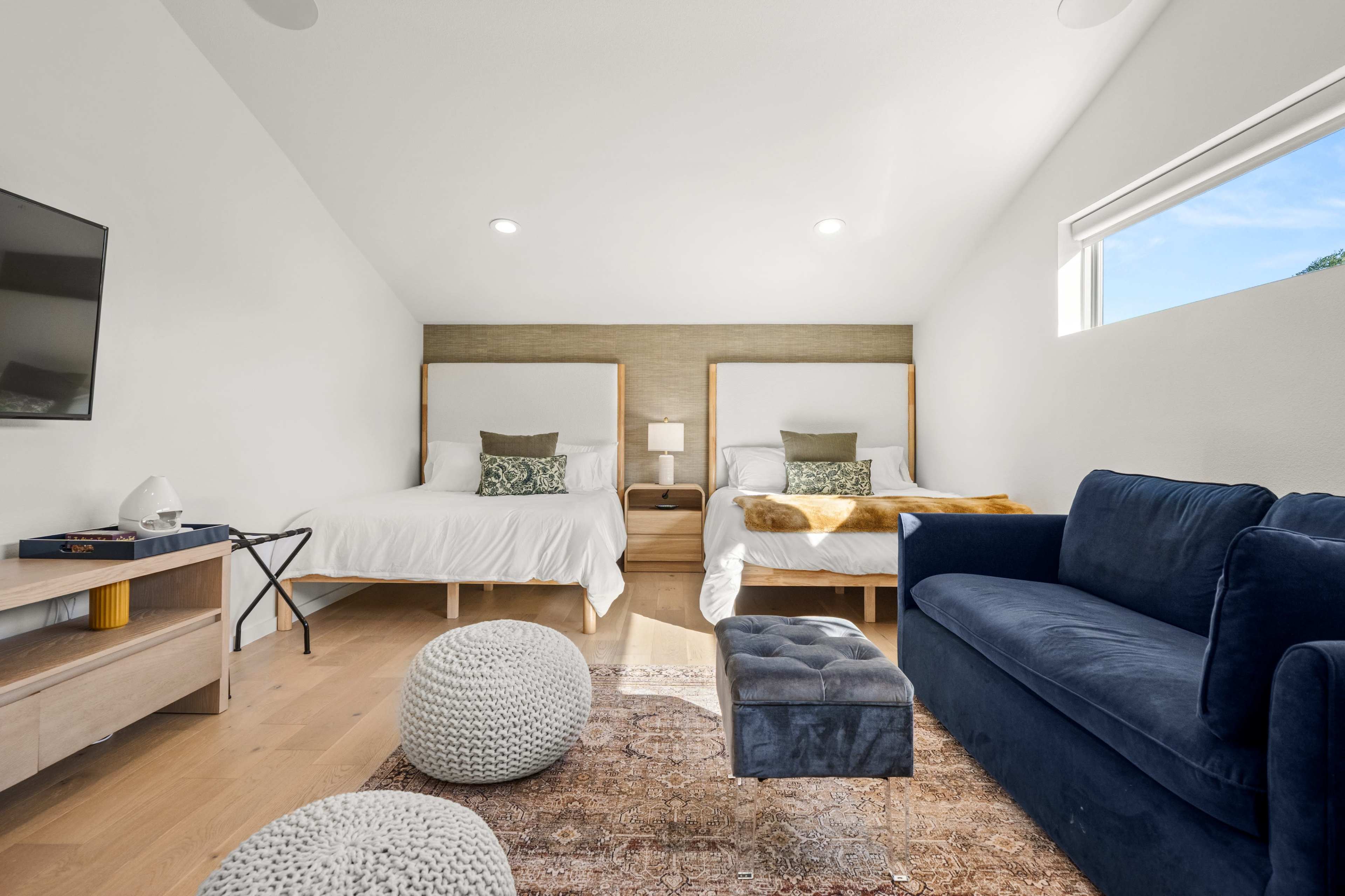 A bright bedroom features two twin beds with wooden headboards, a small nightstand between them, a blue couch, and decorative poufs on a rug.
