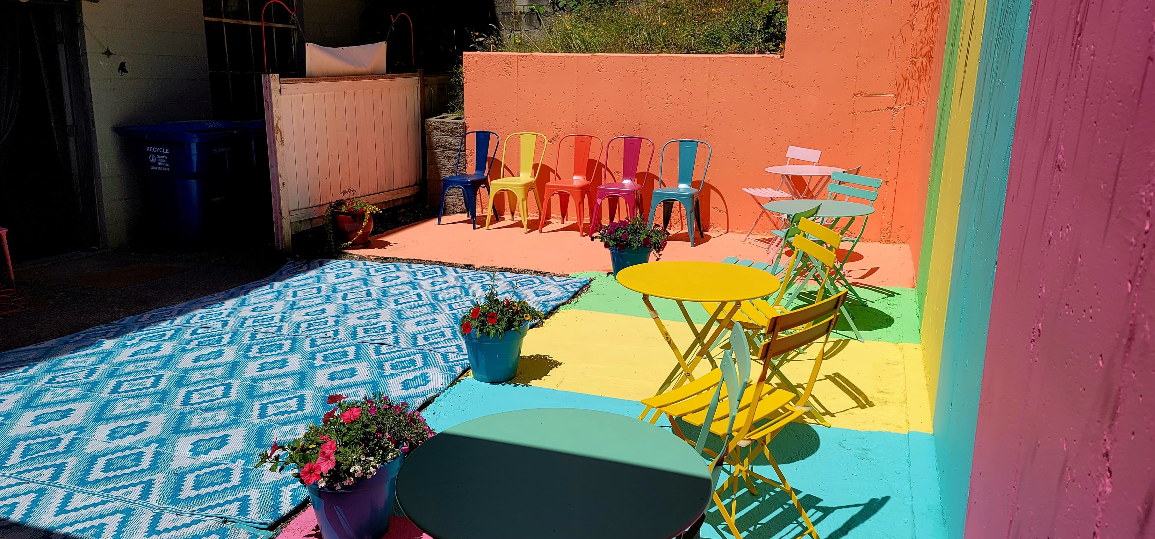 The scene features a vibrant outdoor area with colorful seating, including yellow and teal tables and multicolored chairs, surrounded by potted flowers and a patterned blue rug.