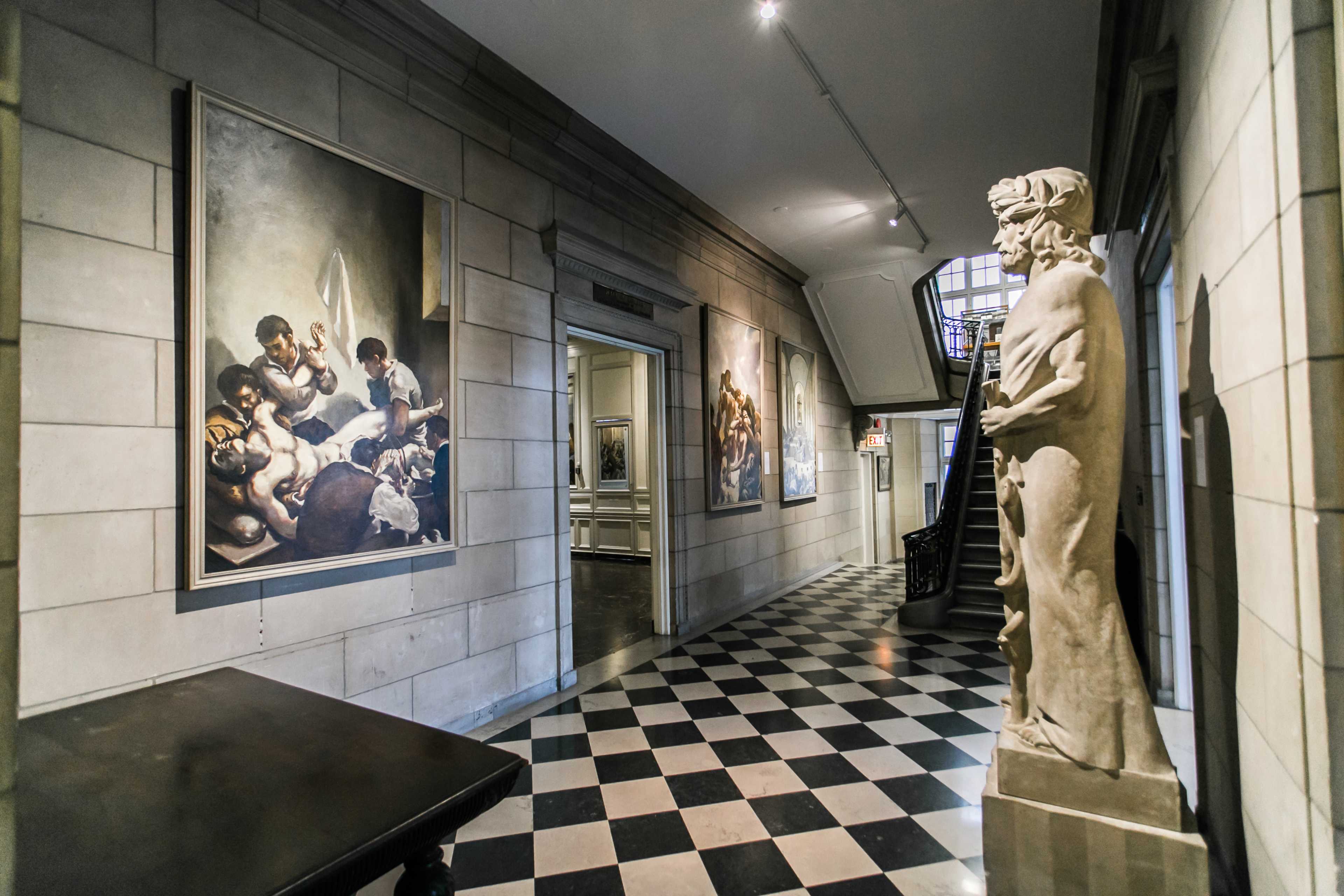 A corridor in an art gallery featuring large paintings on the walls and a statue on the side, with a checkered floor and staircase visible in the background.