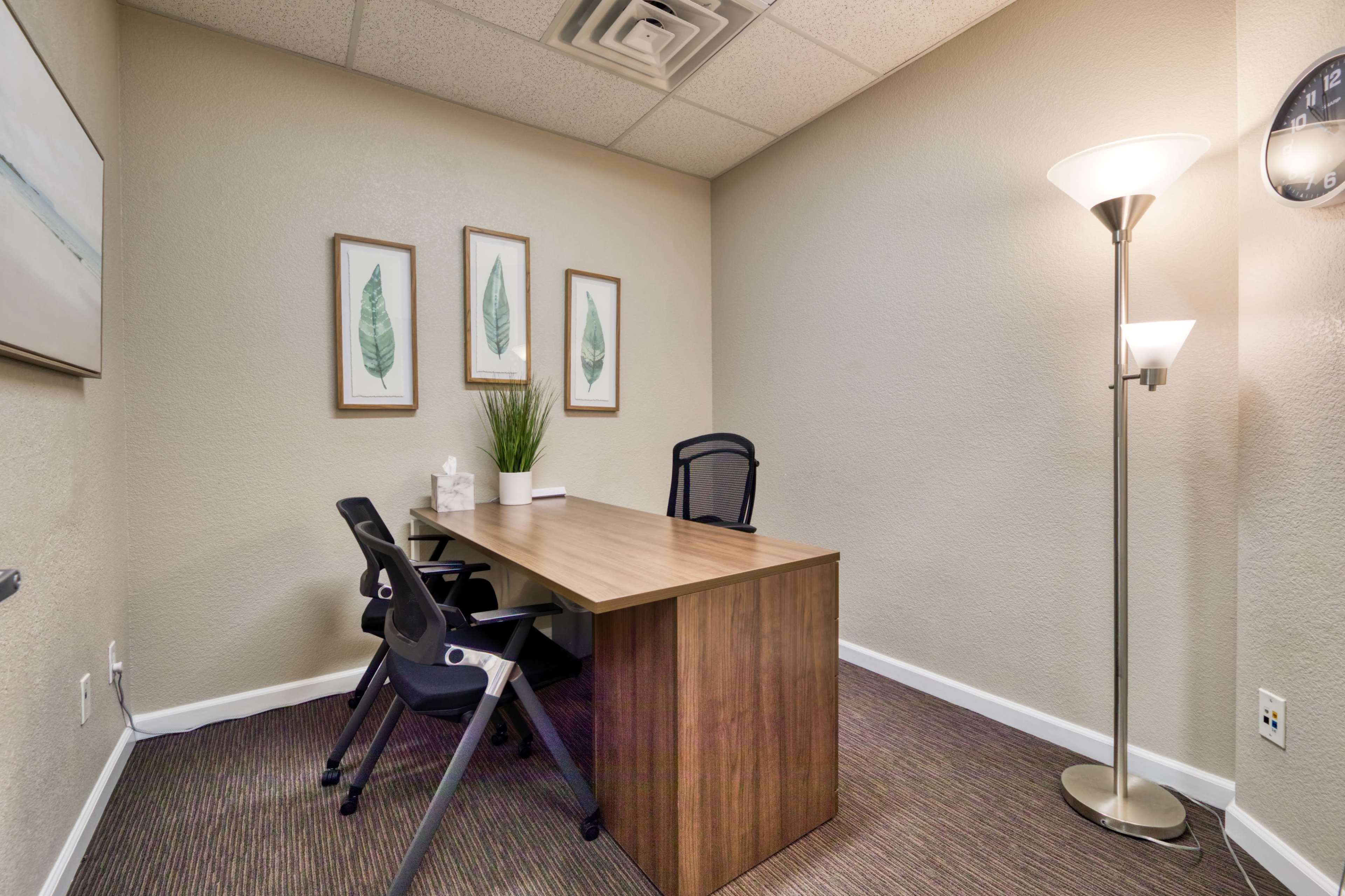 The image shows a small office space featuring a wooden desk, two black chairs, framed leaf artwork on the walls, and a floor lamp.