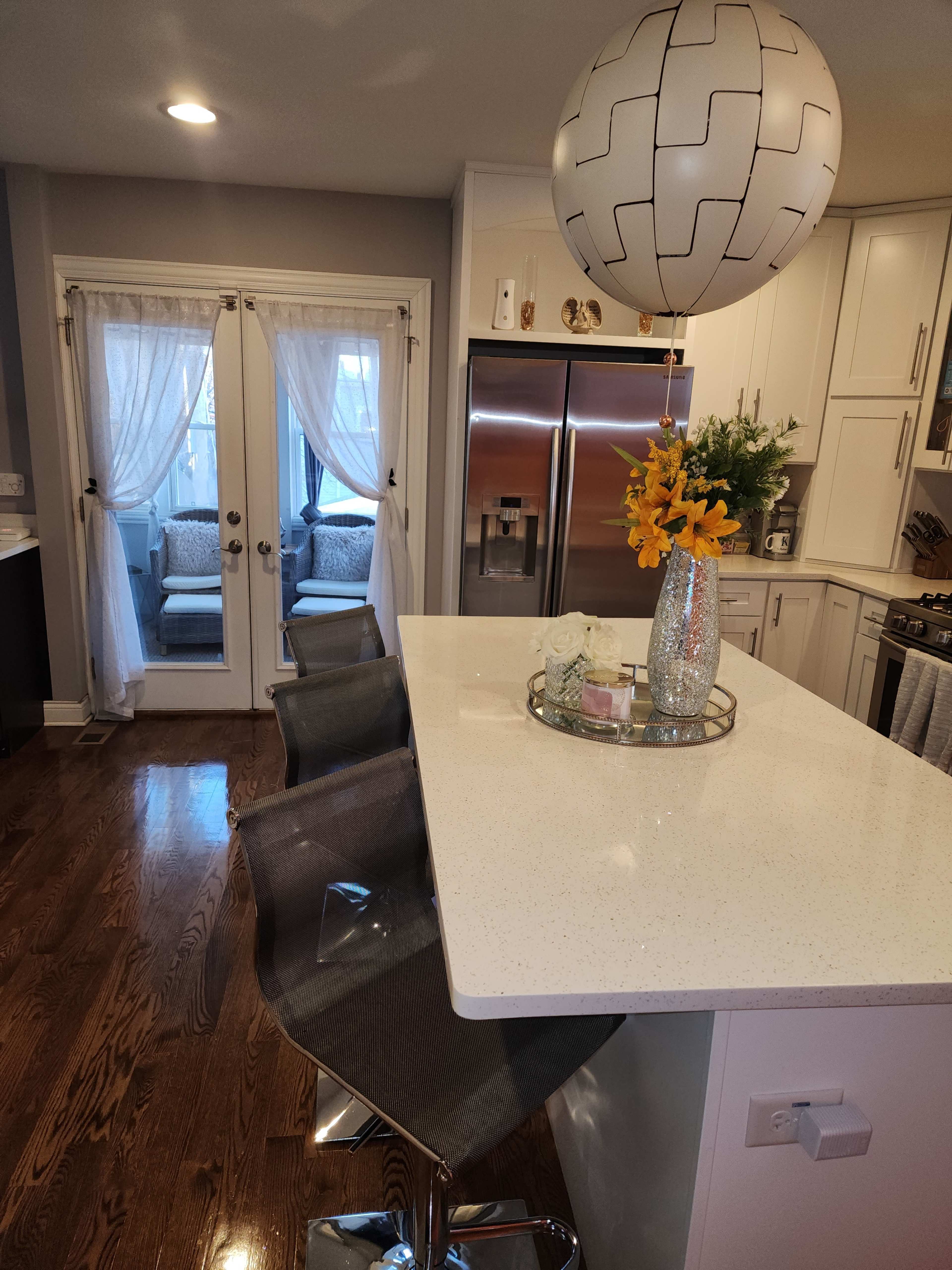A modern kitchen features a bar island with three chairs, a decorative balloon, and a vase of flowers, while sliding glass doors lead to an outdoor seating area.