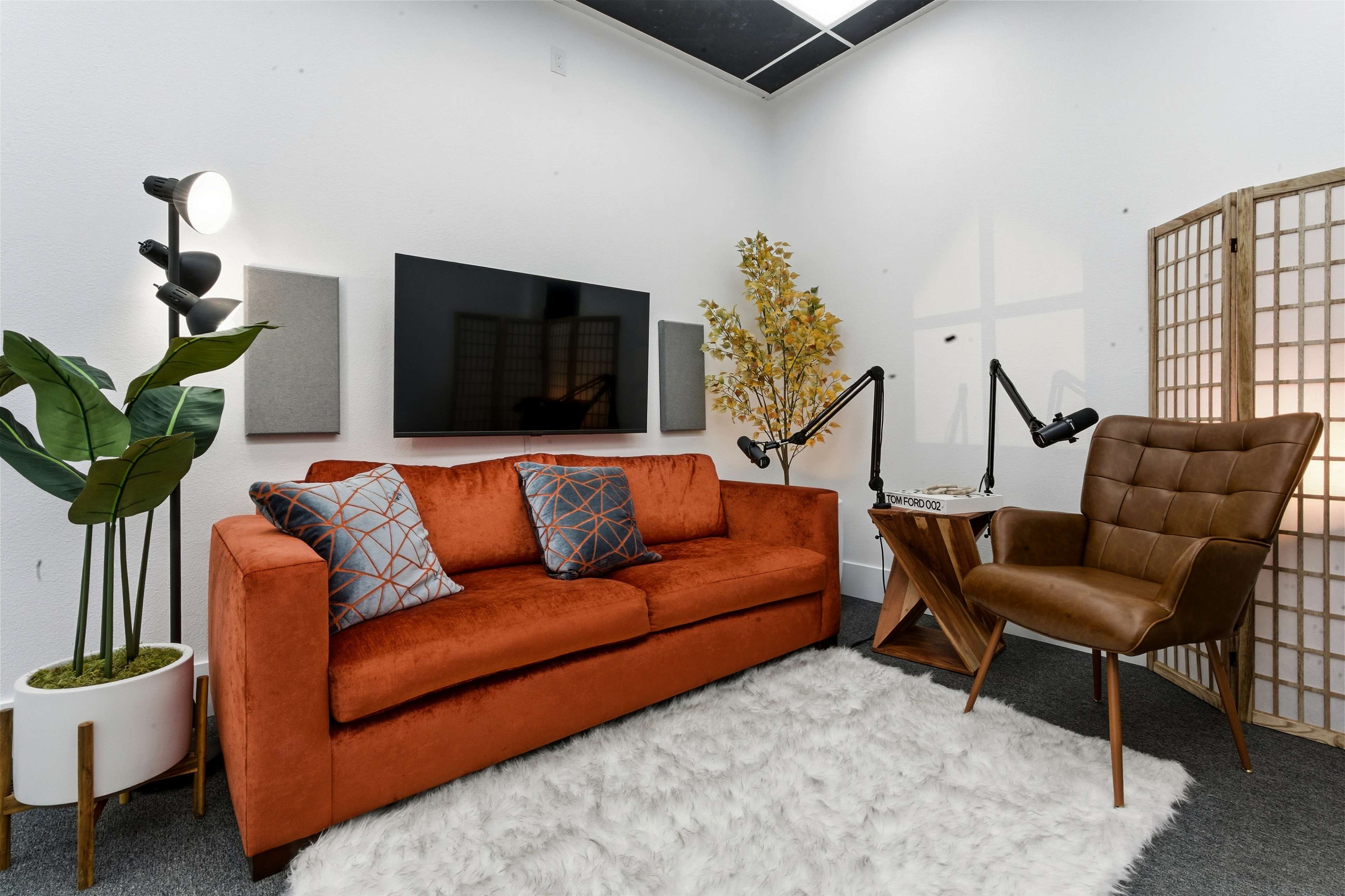 A cozy room features an orange sofa with decorative pillows, a large wall-mounted TV, a potted plant, a wooden chair, and a lighted corner table.