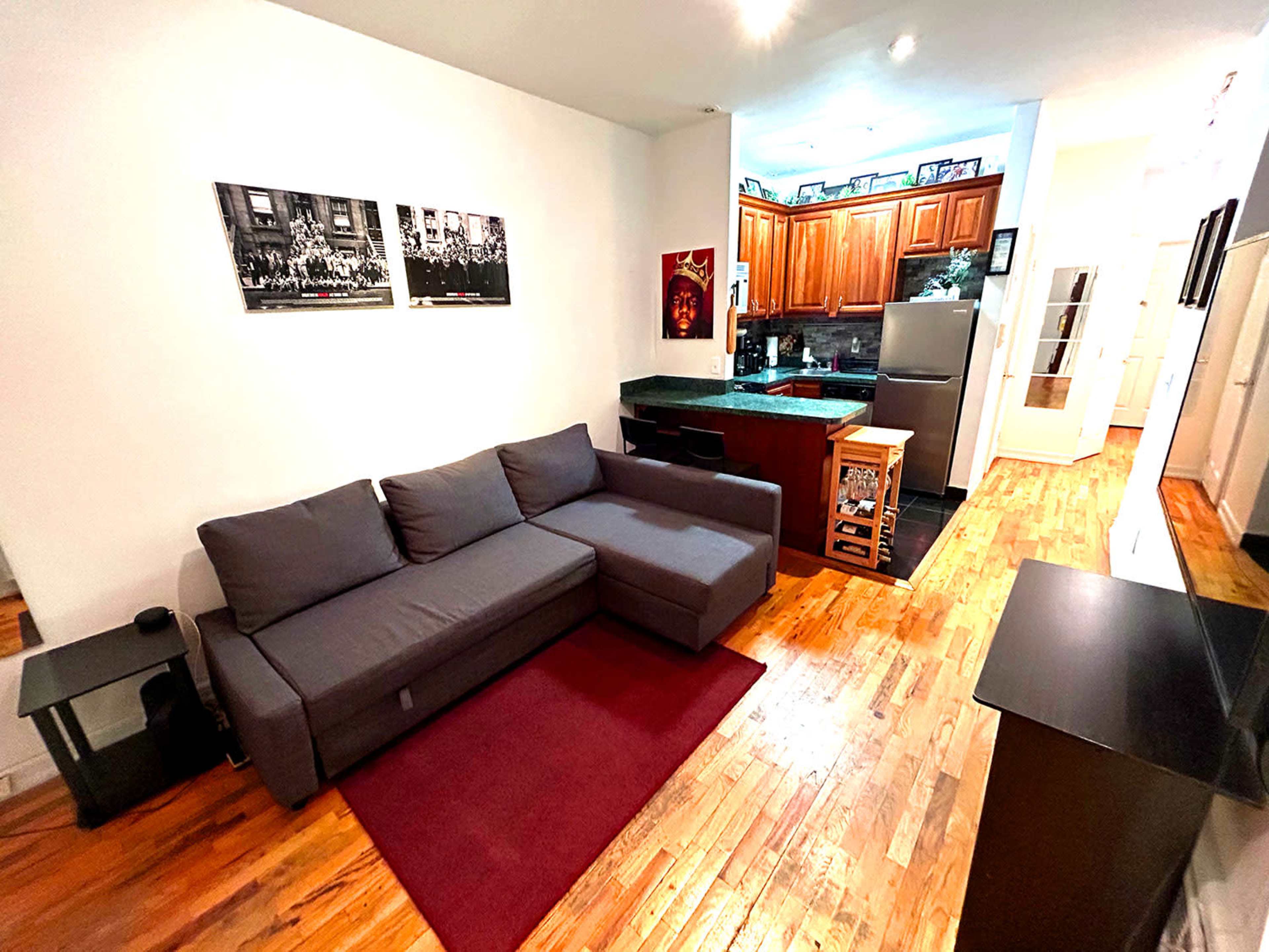 A cozy living space with a gray sectional sofa, a red rug, and a kitchen area featuring wooden cabinets and stainless steel appliances.