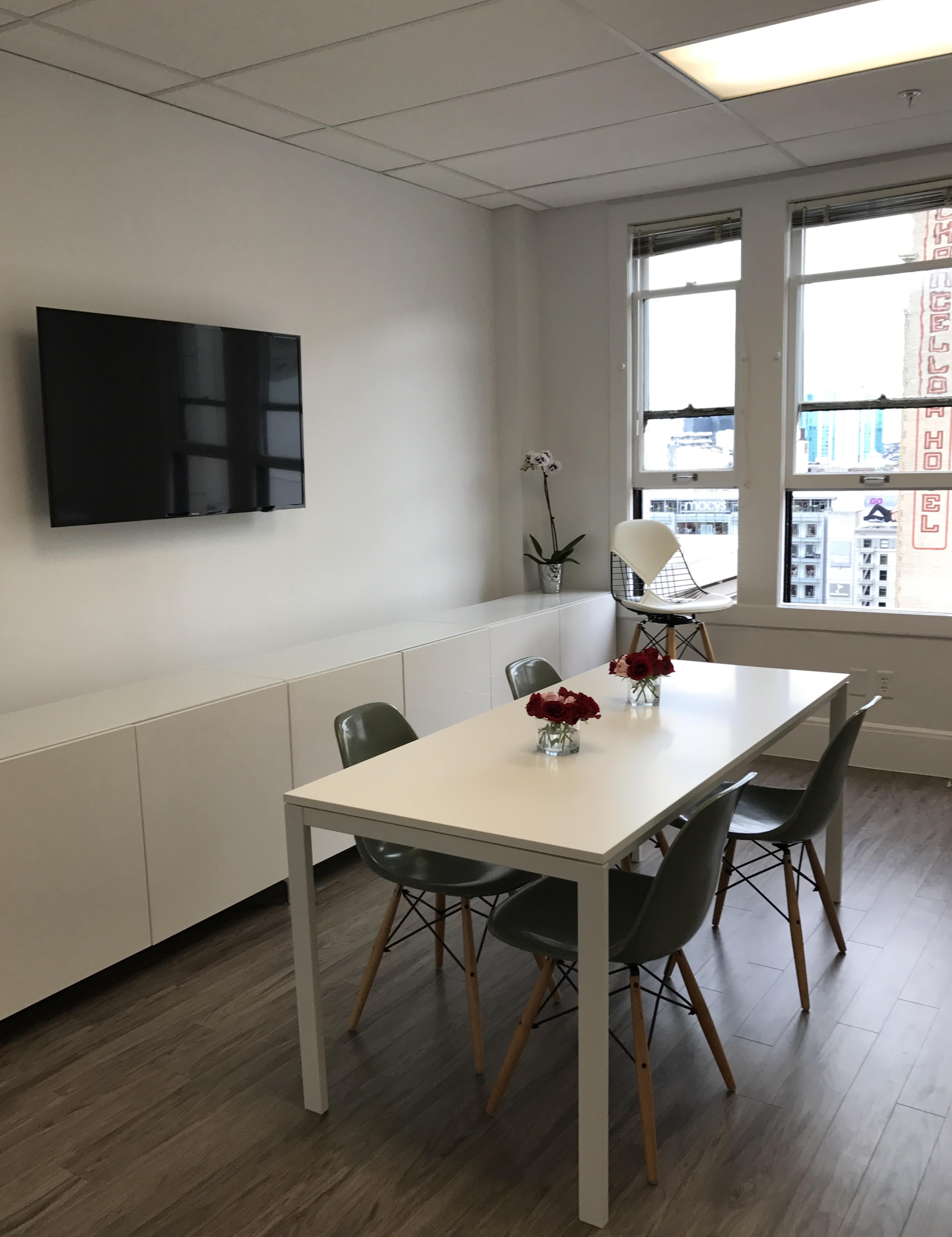 A modern meeting room features a white table surrounded by four green chairs, a wall-mounted television, and large windows with a view of a cityscape.