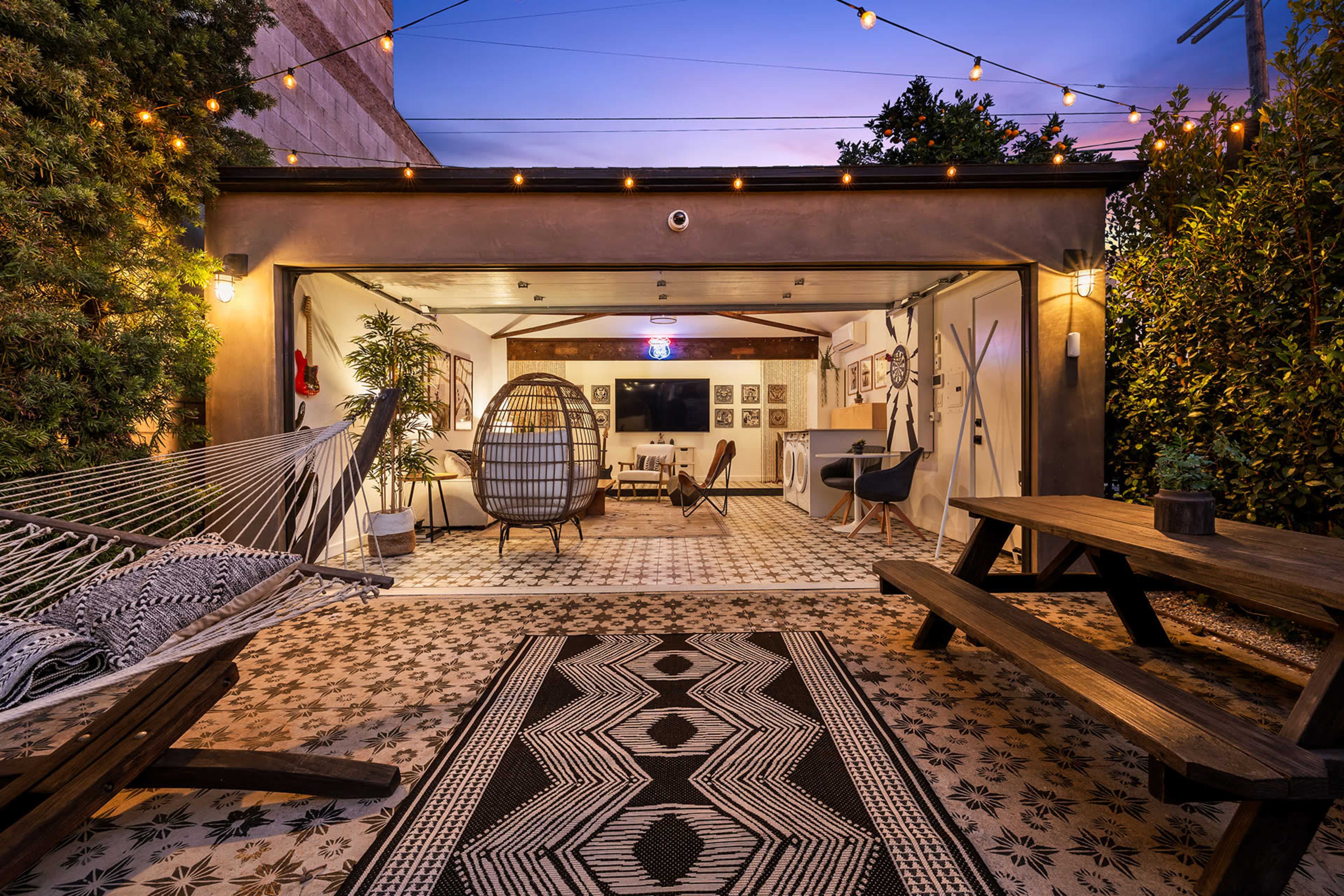 The scene features a cozy outdoor space with a hammock, a hanging chair, a picnic table, and decorative string lights under a twilight sky.