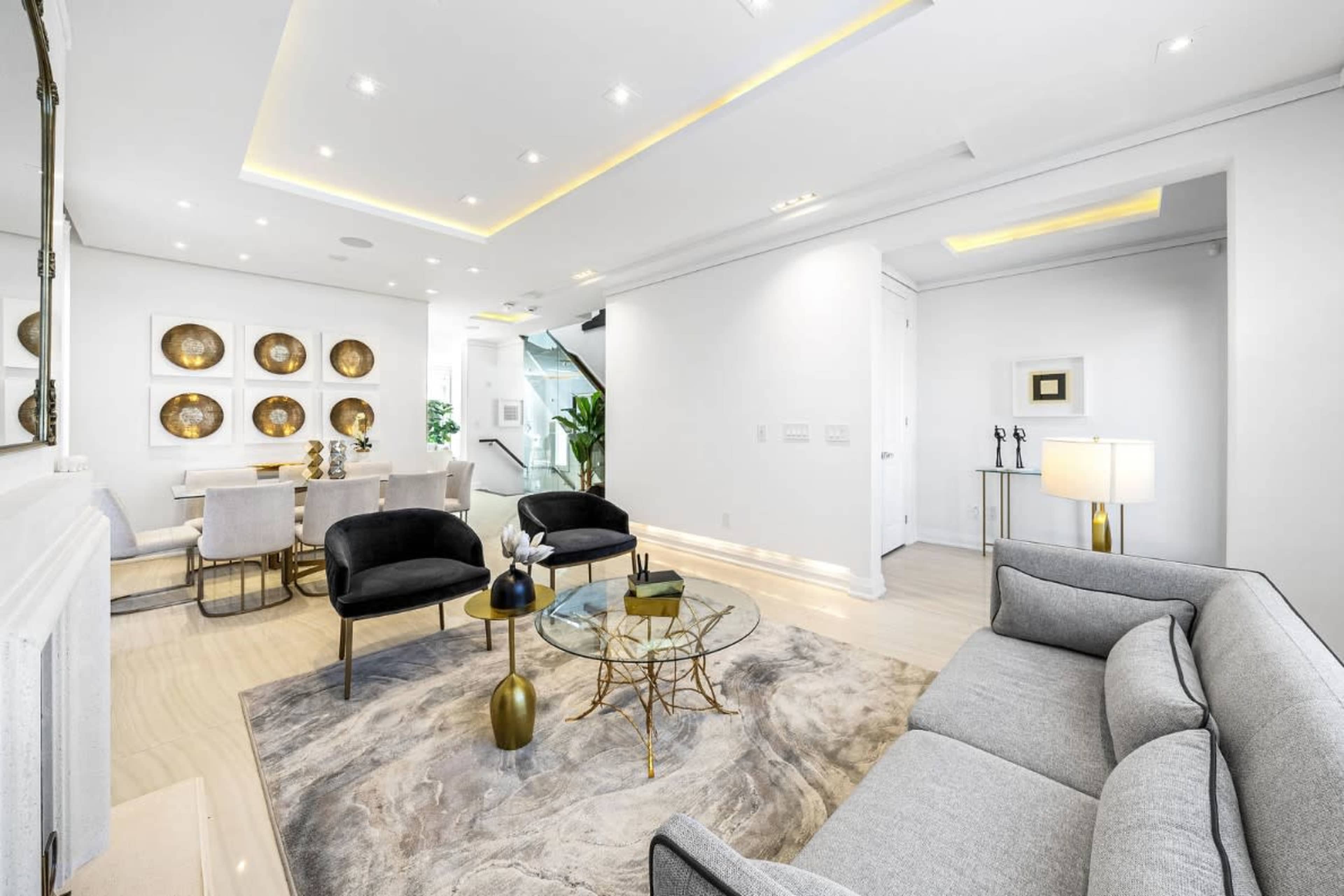 The image shows a modern, well-lit living room featuring a gray sofa, a glass coffee table, and a dining area with a round table and decorative gold plates on the wall.