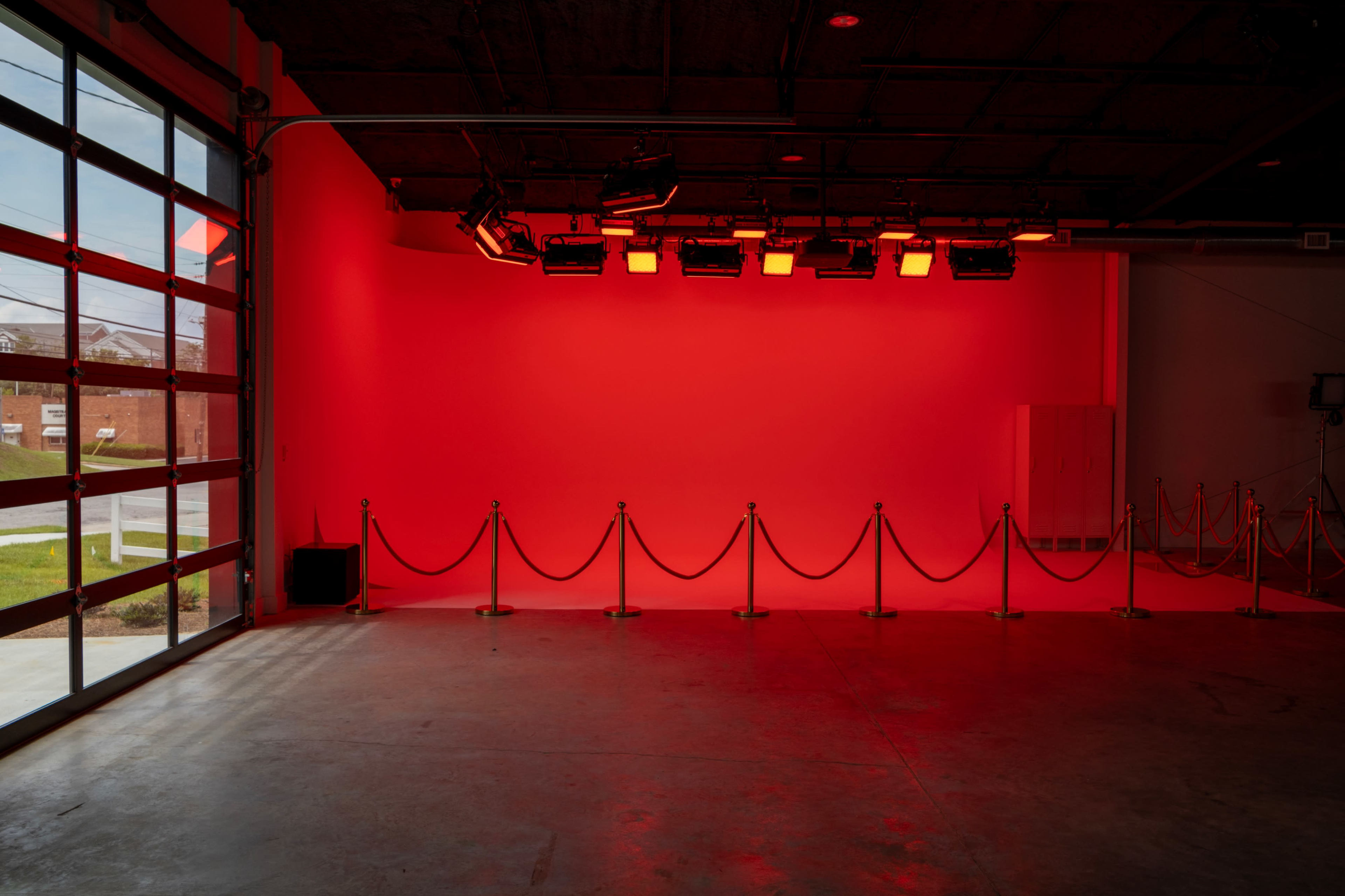 The image shows an empty studio space with a red lit wall, featuring lighting equipment overhead and a velvet rope barrier in front.