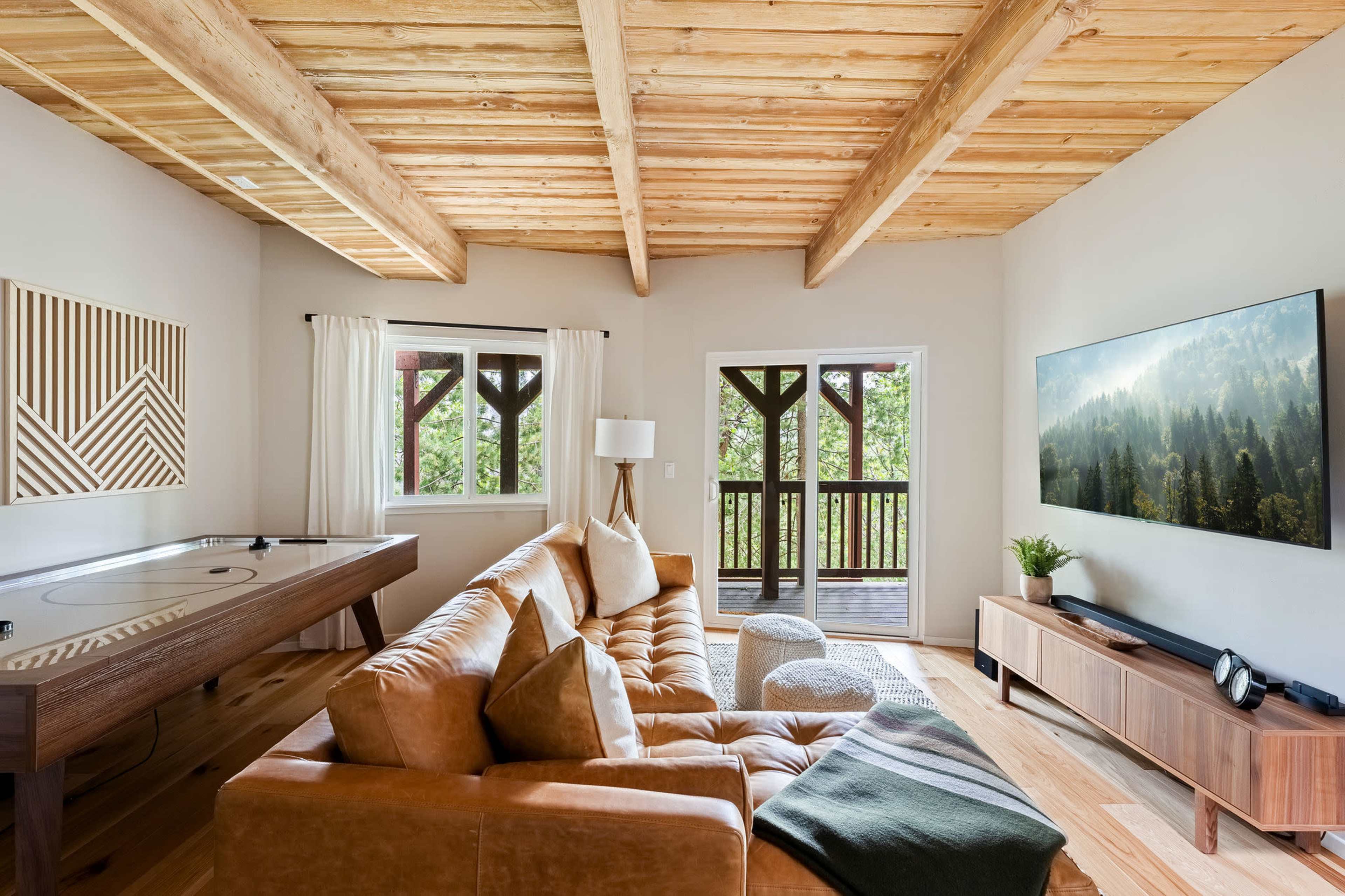 The living room features a large leather sectional couch, a wooden table, and a wall-mounted television, with large windows providing a view of the outdoors.