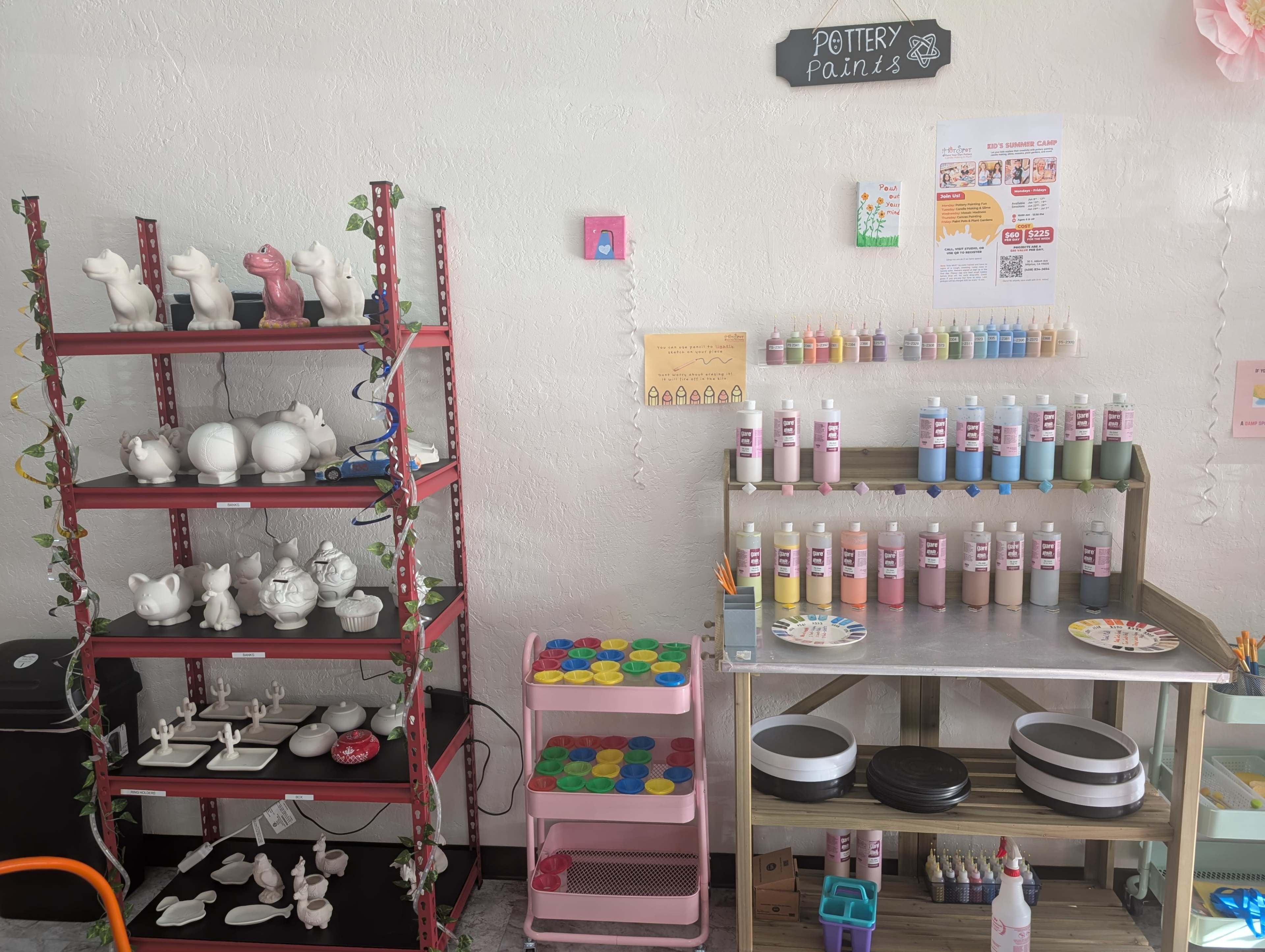 The image shows a brightly lit pottery painting studio with shelves displaying unpainted ceramic items and a variety of paint bottles organized by color.