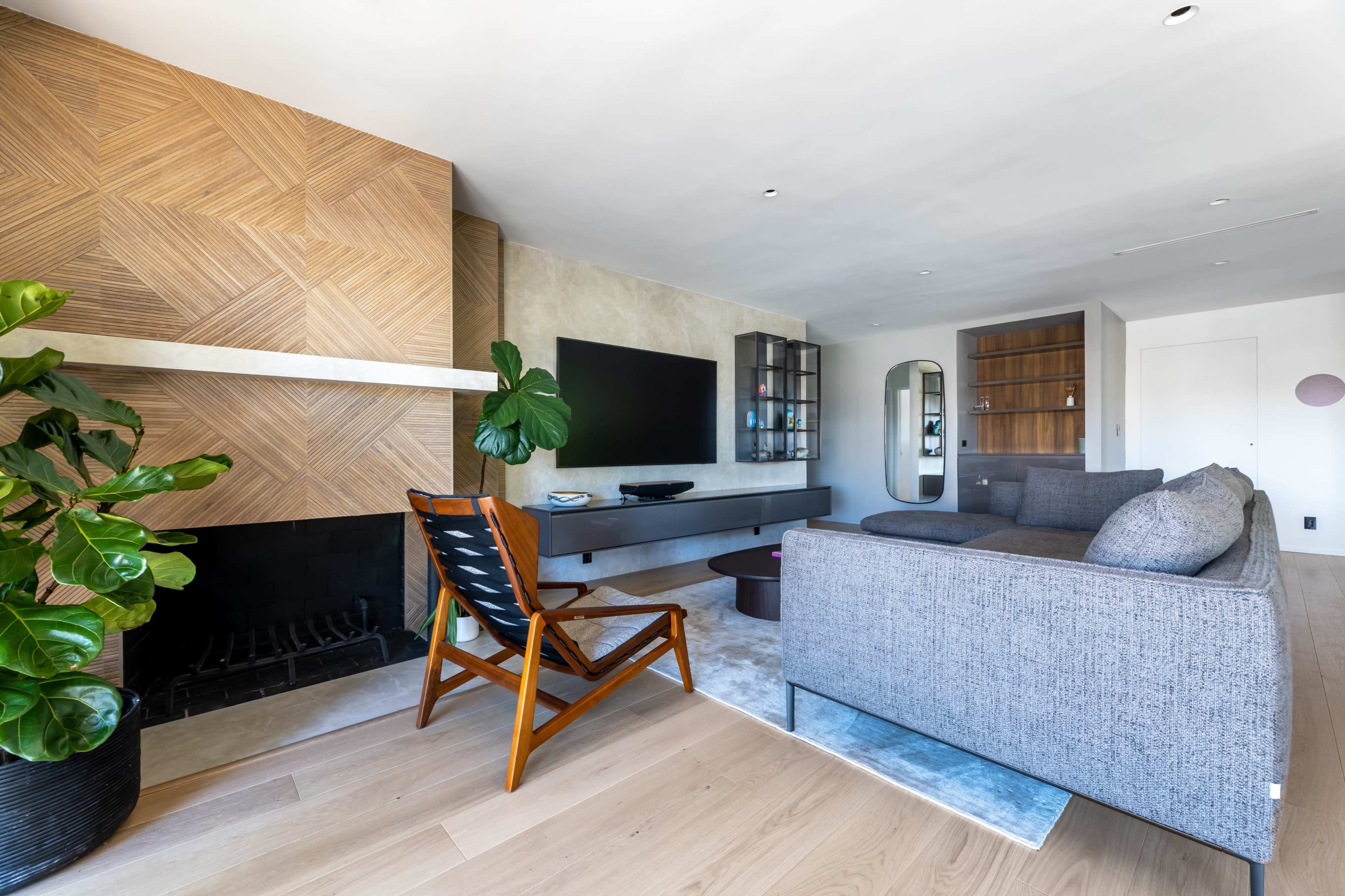 A modern living room featuring a patterned wood accent wall, a flat-screen TV, a gray sectional sofa, and a wooden chair next to a fireplace.