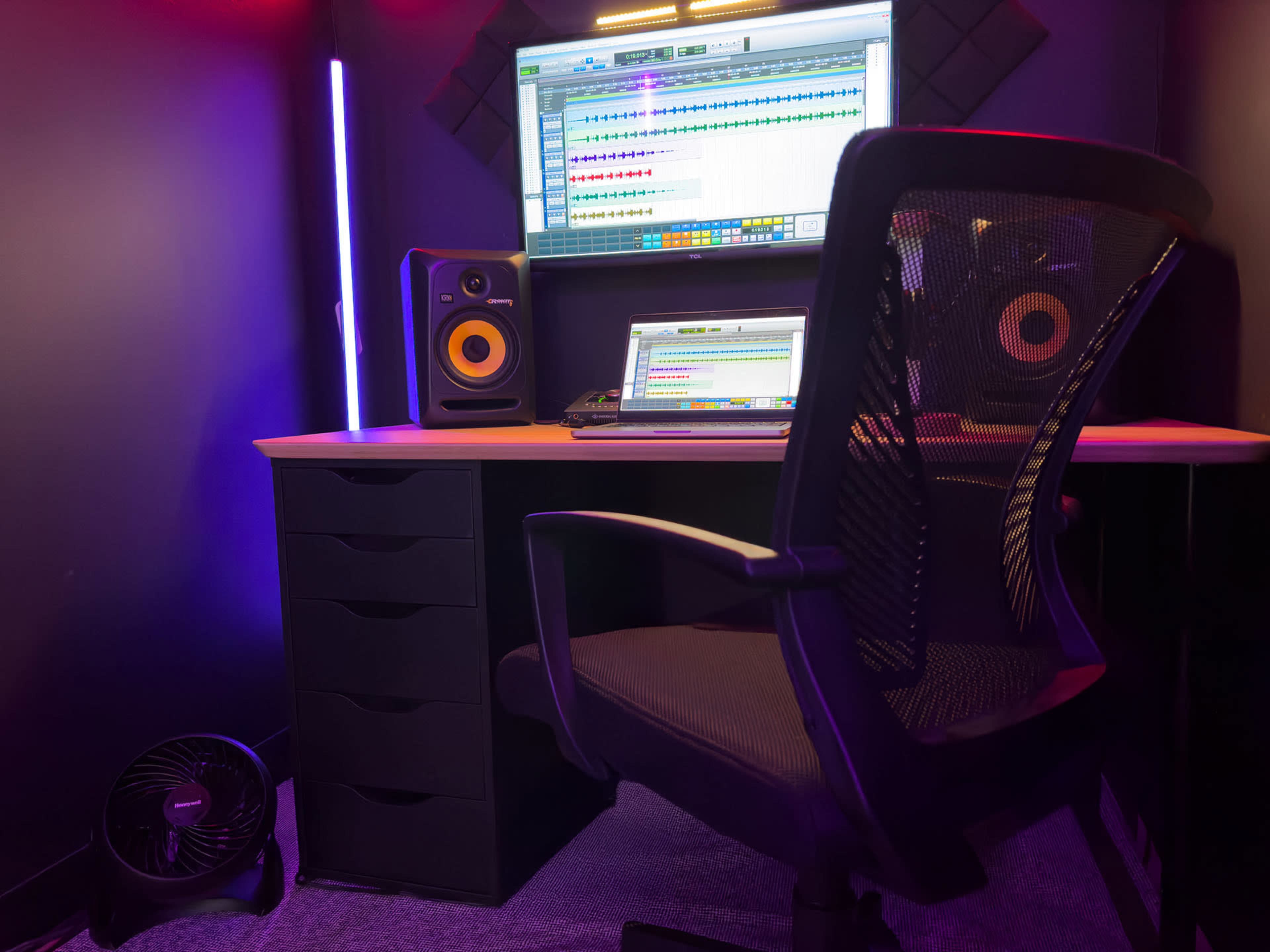 The image shows a recording studio setup with a desk, a computer monitor displaying audio tracks, studio monitors, and an office chair.