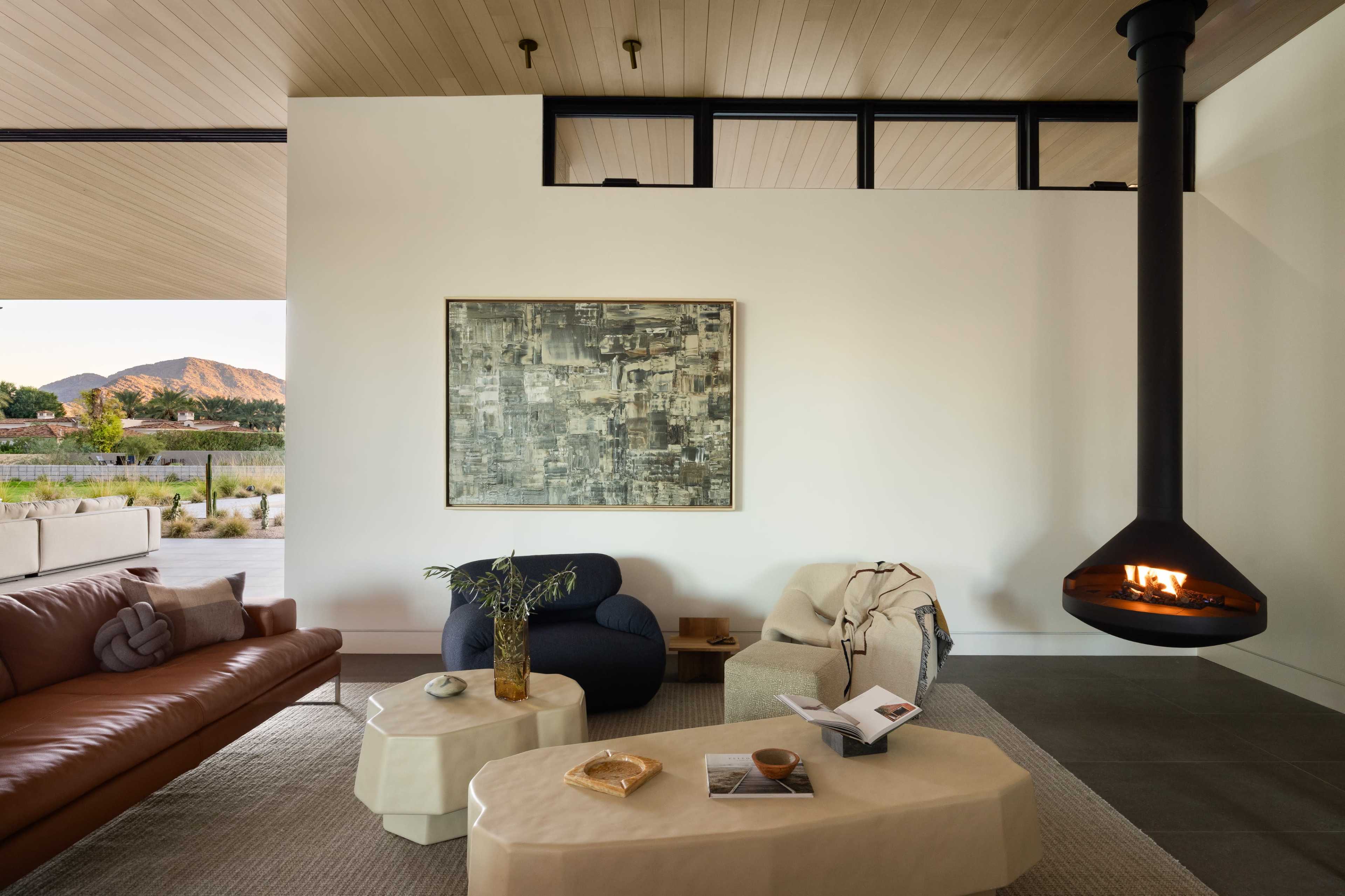 The image shows a modern living room featuring a brown leather sofa, blue and beige chairs, a minimalist coffee table, and a suspended fireplace, with large windows showcasing a mountainous landscape outside.