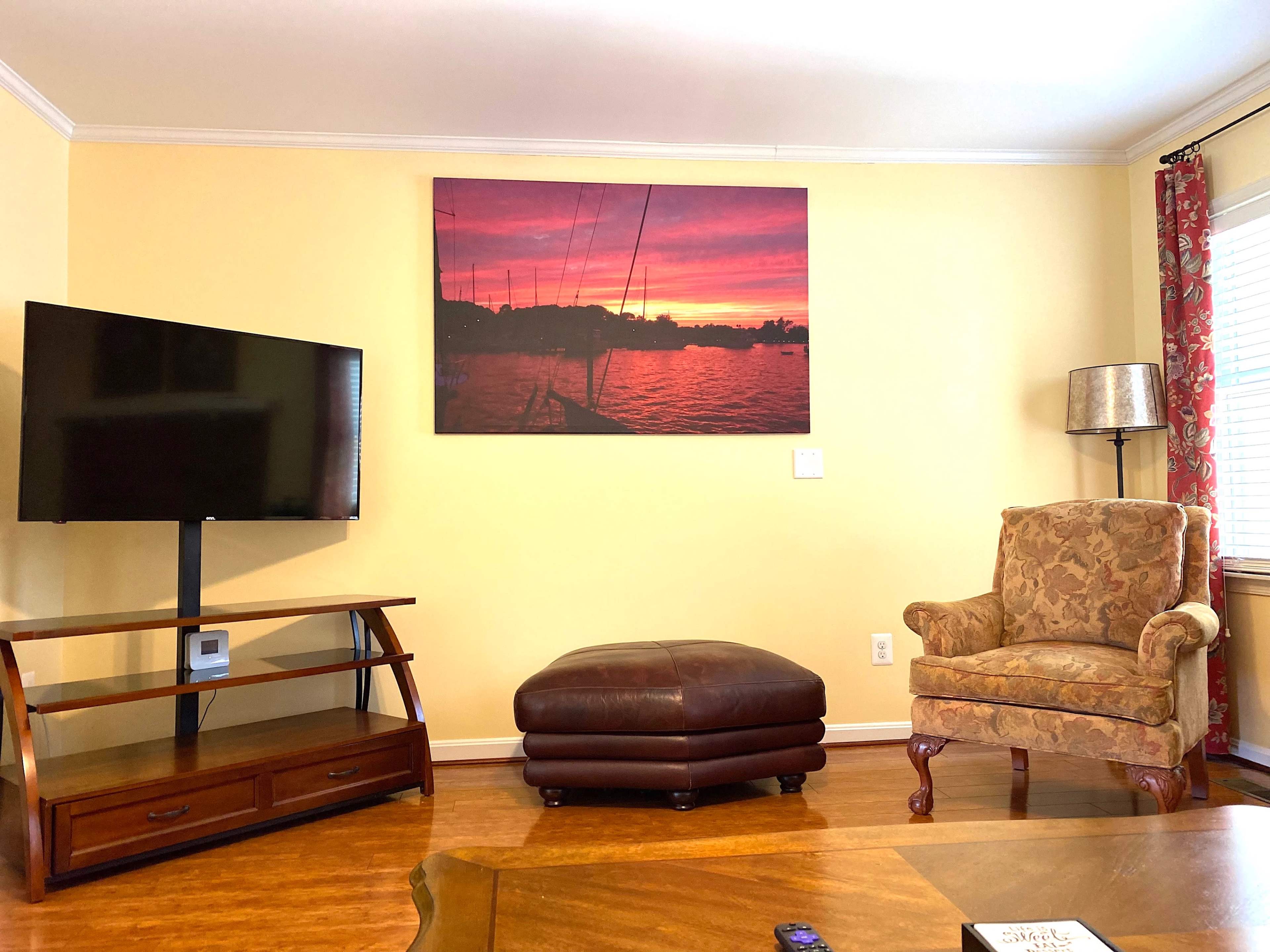 A living room features a television mounted on the wall, a large sunset photograph, a brown ottoman, and an armchair beside a lamp.