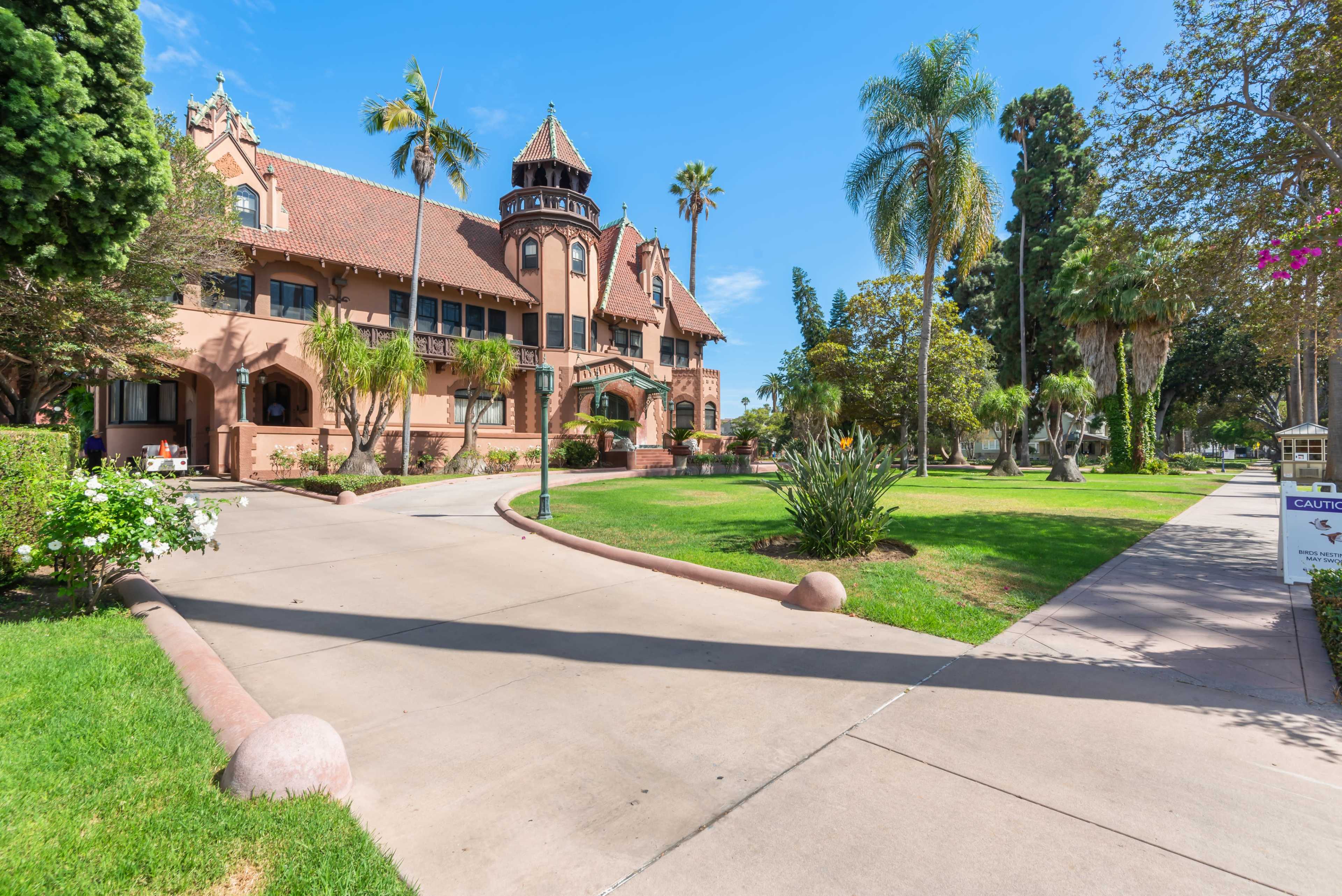 The image shows a large, historic building with a tower, surrounded by palm trees and well-maintained grassy areas along a pathway.
