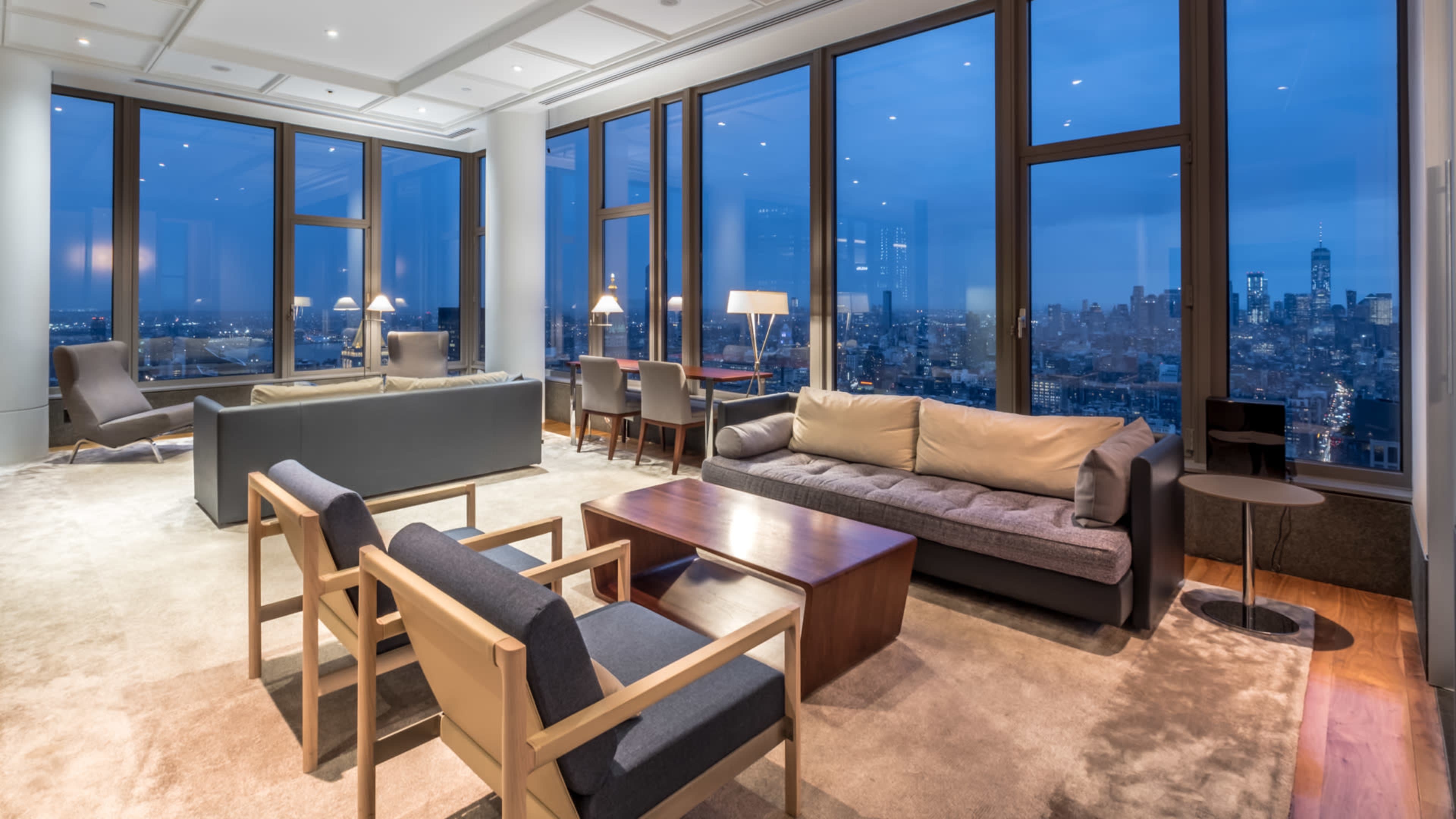 A modern living room with large windows offering a view of a city skyline at dusk, featuring minimalist furniture and soft lighting.