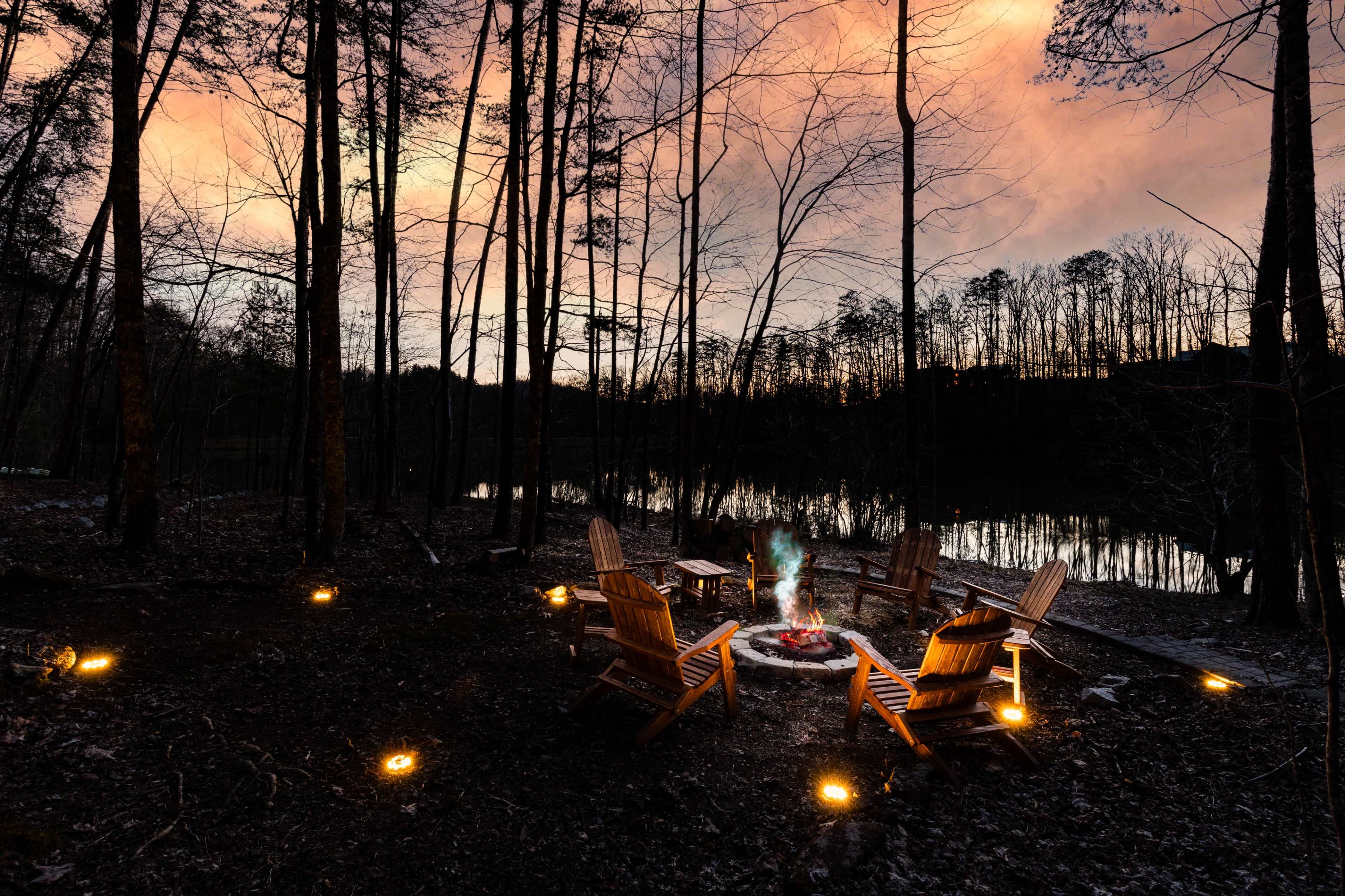 A circle of wooden chairs surrounds a fire pit near a calm lake, with trees silhouetted against a colorful sunset sky.