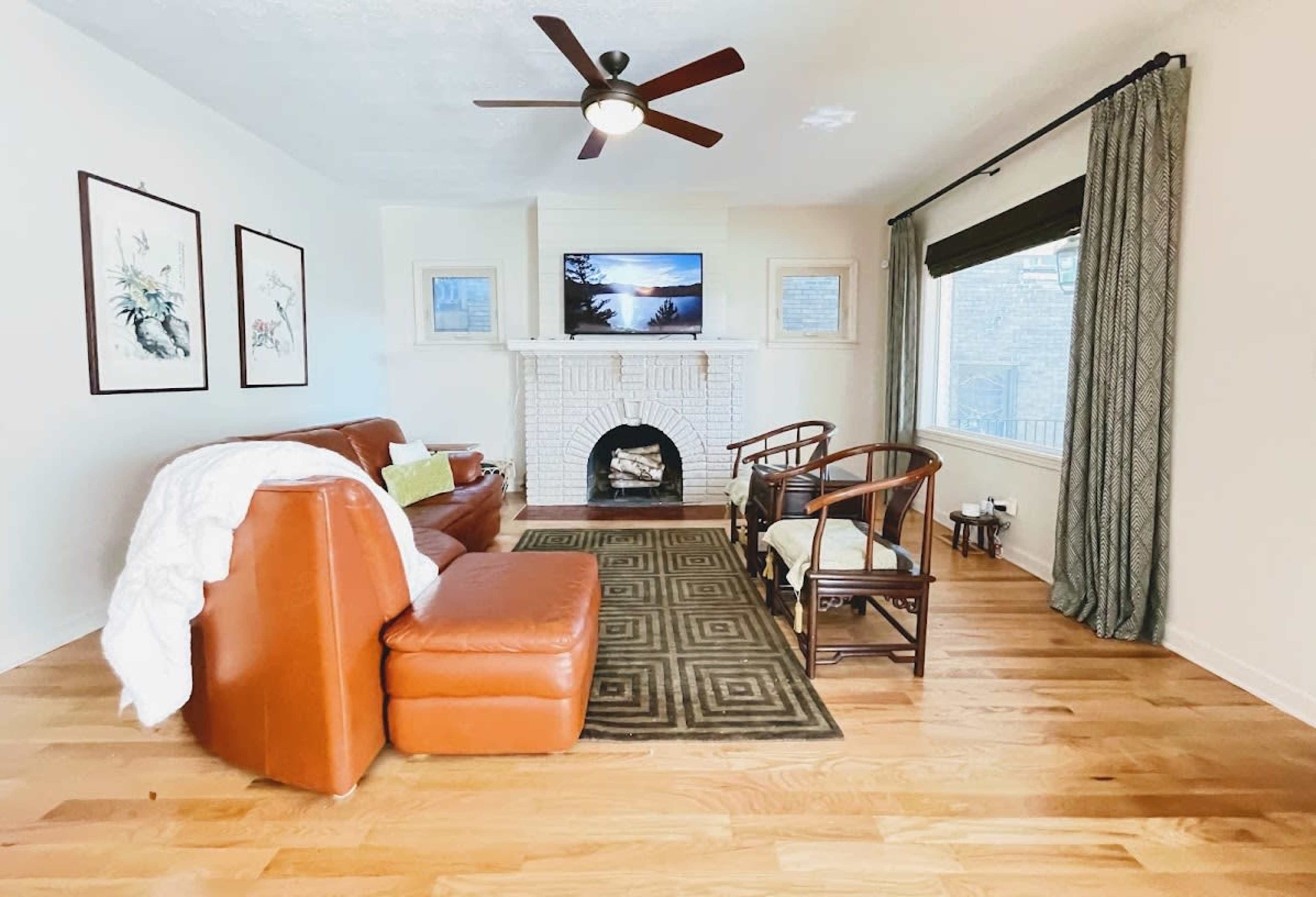 A living room features a brown leather sofa, two wooden chairs, a white brick fireplace, and a television mounted on the wall.
