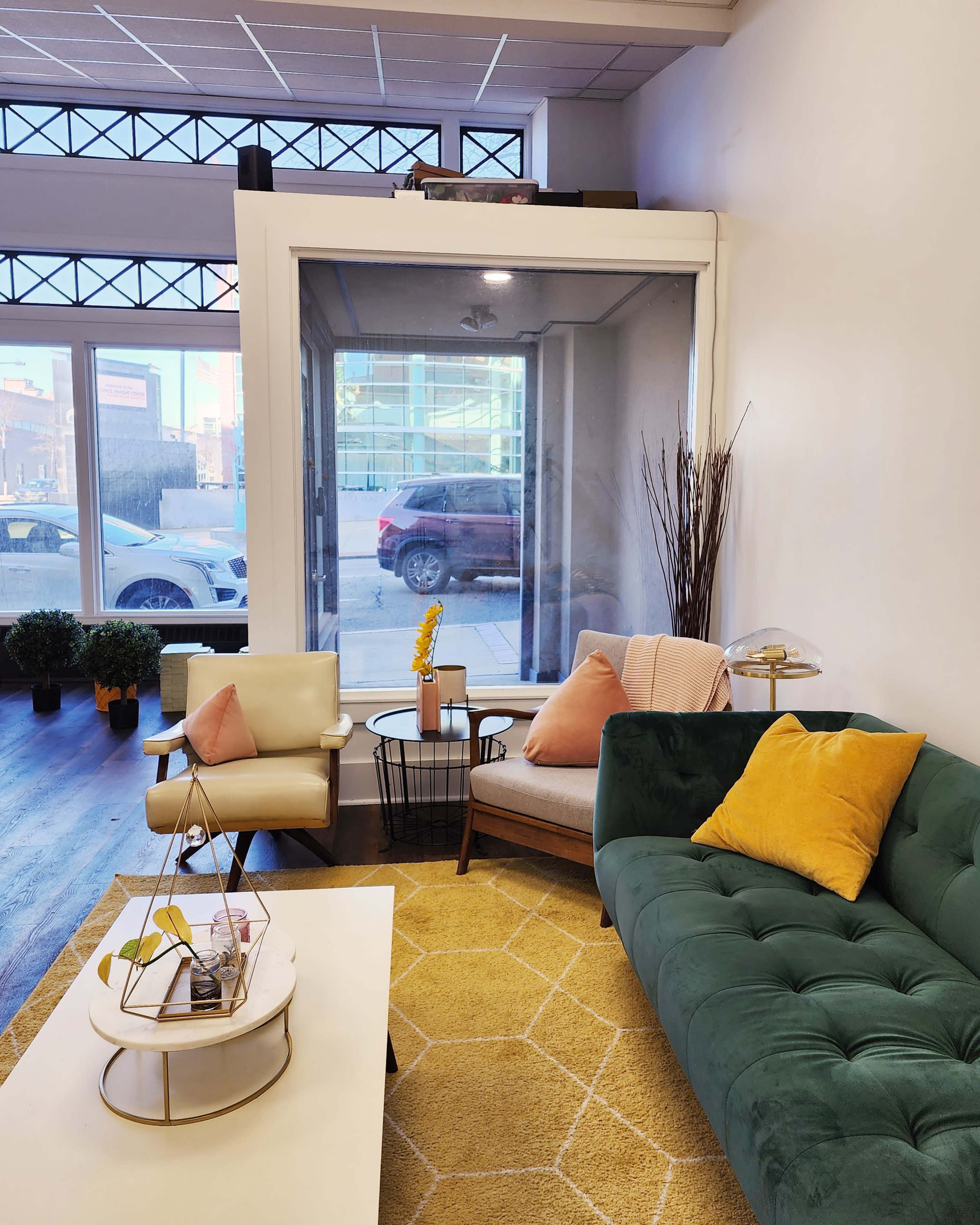 The scene features a modern lounge area with a green sofa, a beige armchair, a coffee table, and large windows revealing a city street outside.