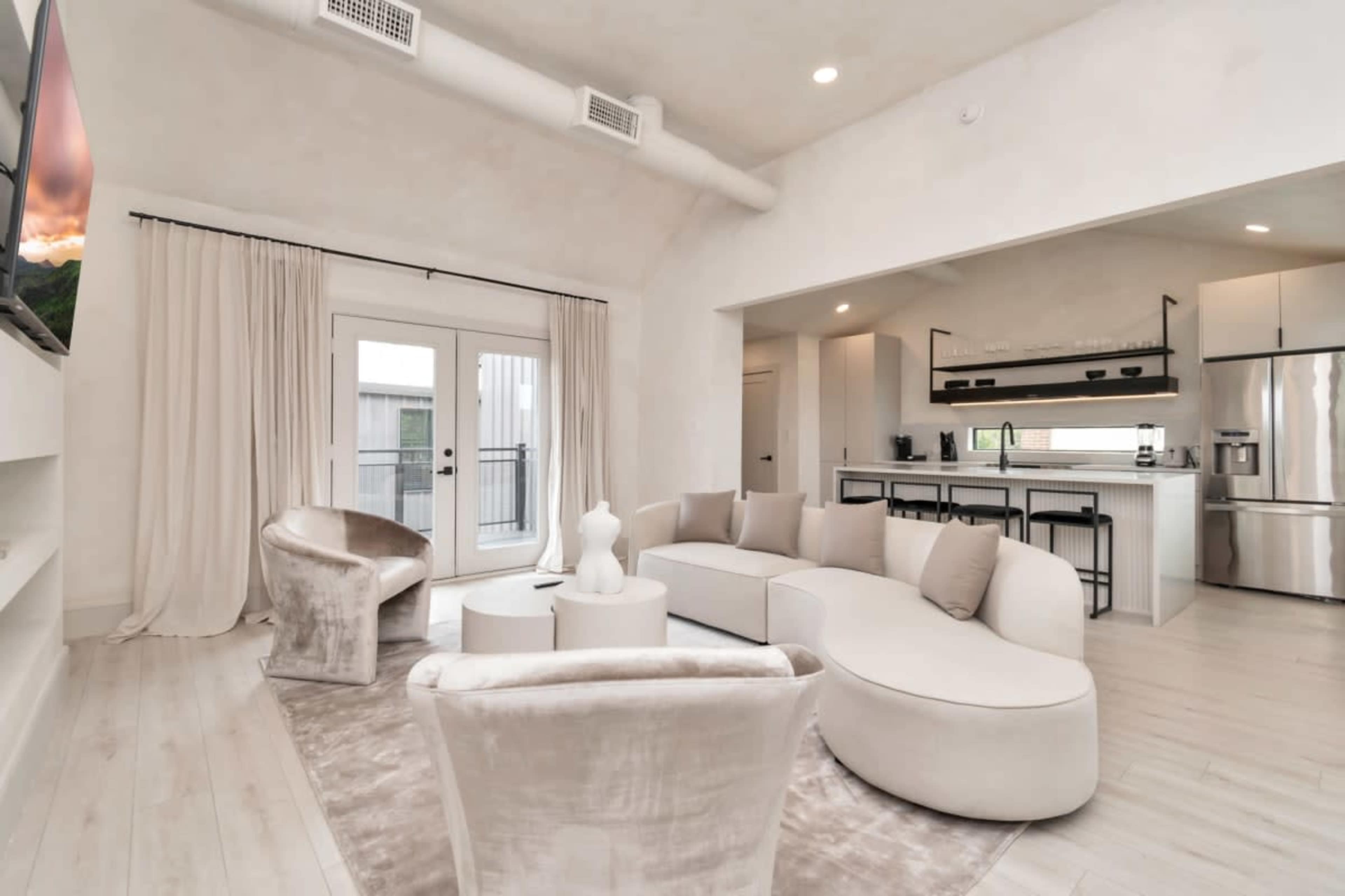 A modern living space features a curved sectional sofa, two armchairs, and a kitchenette with stainless steel appliances, all set against light-colored walls and flooring.