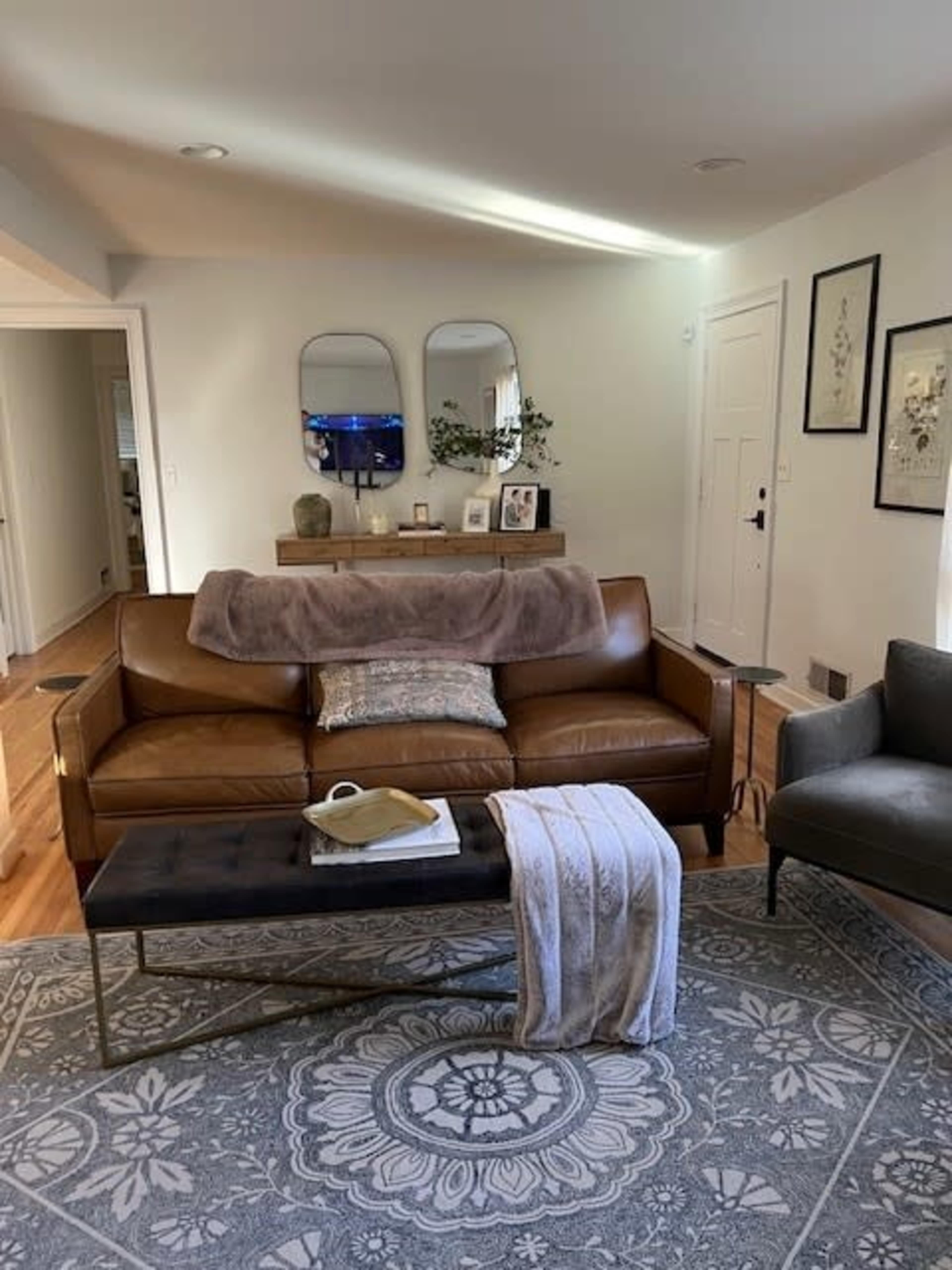 A brown leather sofa with a throw blanket is positioned in a living room adorned with decorative mirrors, artwork, and a coffee table on a patterned rug.