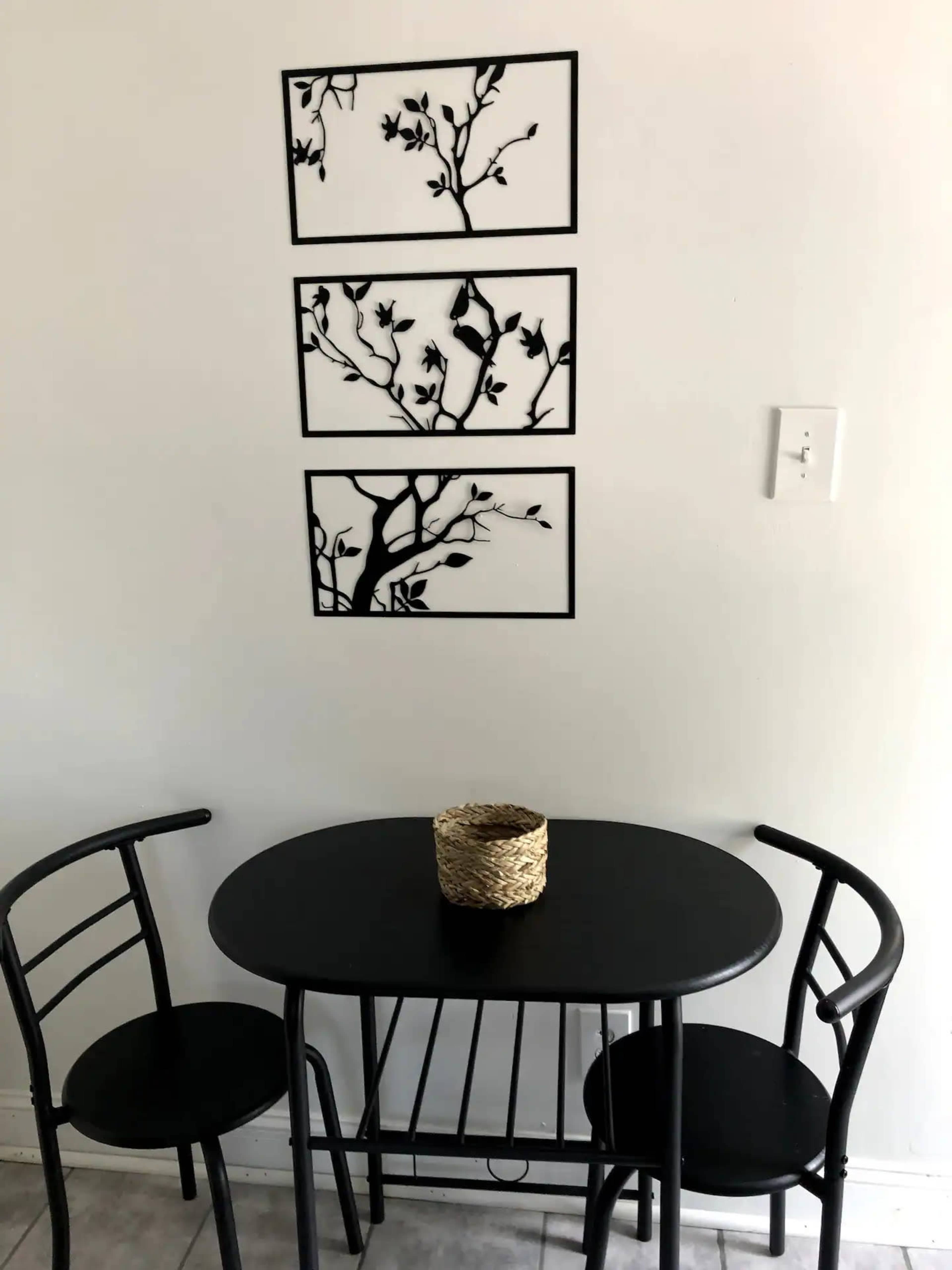 A small black table with two chairs is positioned in front of a white wall featuring three framed black wall decorations of branches and leaves.