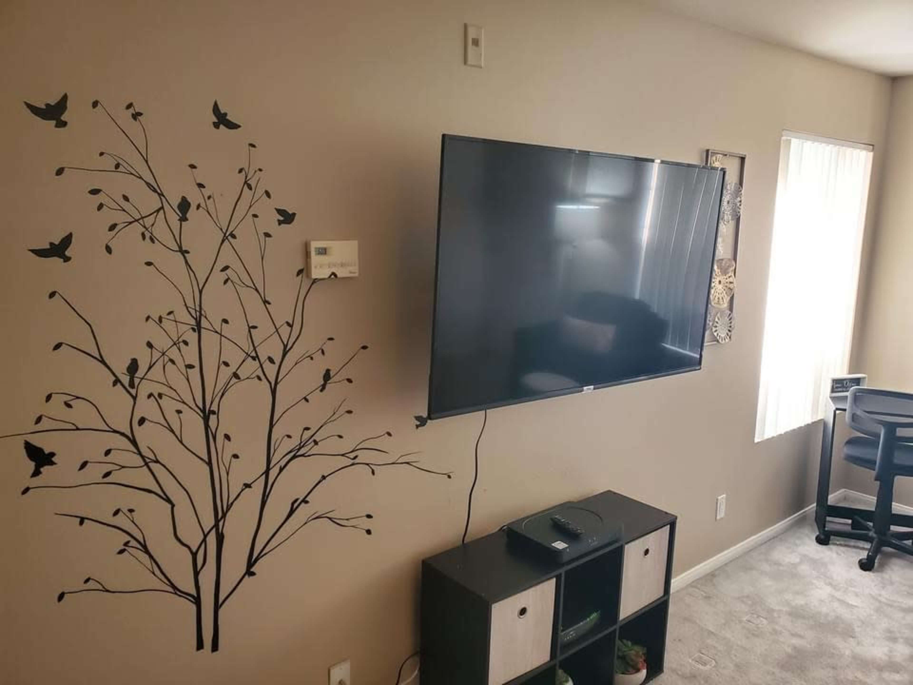 A television is mounted on a wall next to a decorative tree branch decal, with a small shelf unit below it and a desk visible in the background.