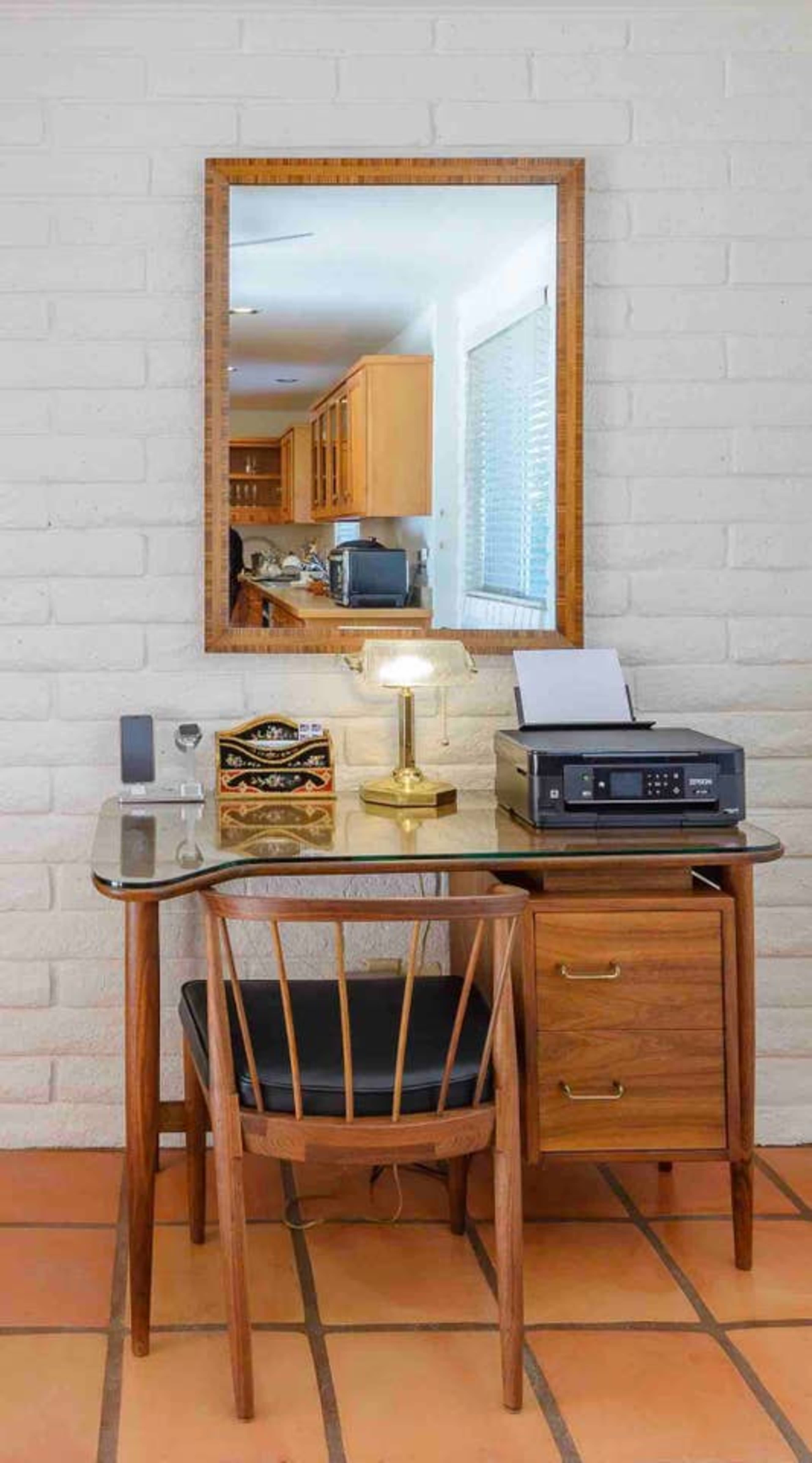 A wooden desk with a printer, lamp, and drawer is placed against a white brick wall, reflecting a kitchen area in a nearby mirror.