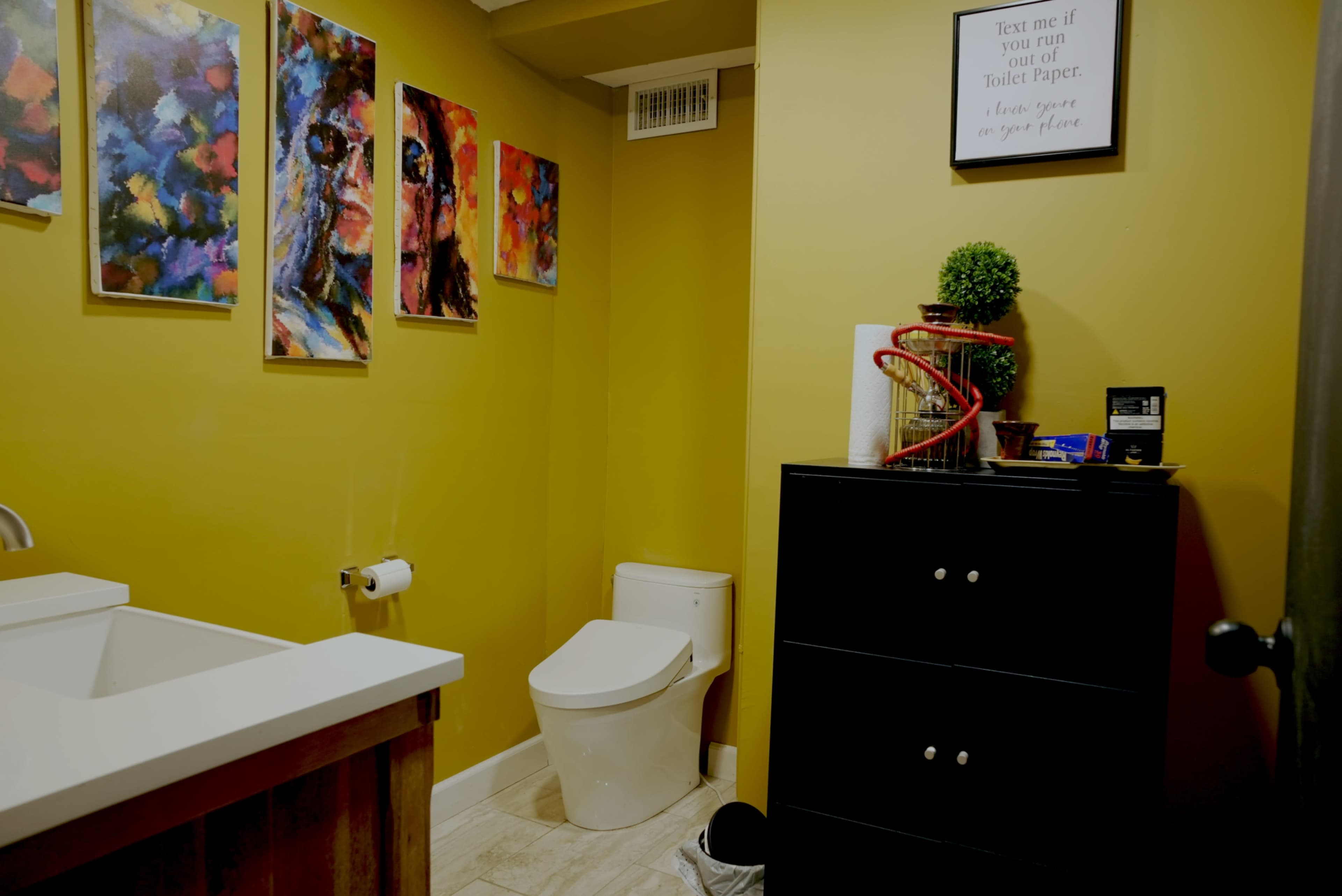 A small bathroom with a beige countertop sink, a white toilet, and colorful artwork on the yellow walls.