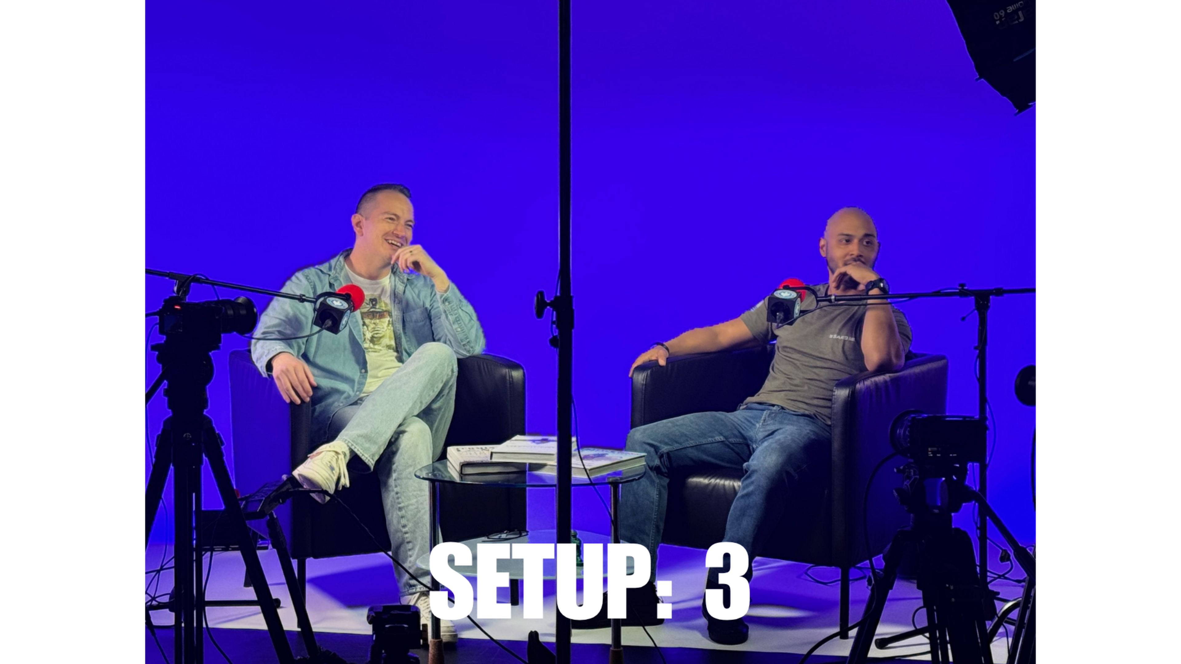 Two men sit on black chairs facing each other, surrounded by cameras and studio equipment against a blue backdrop.