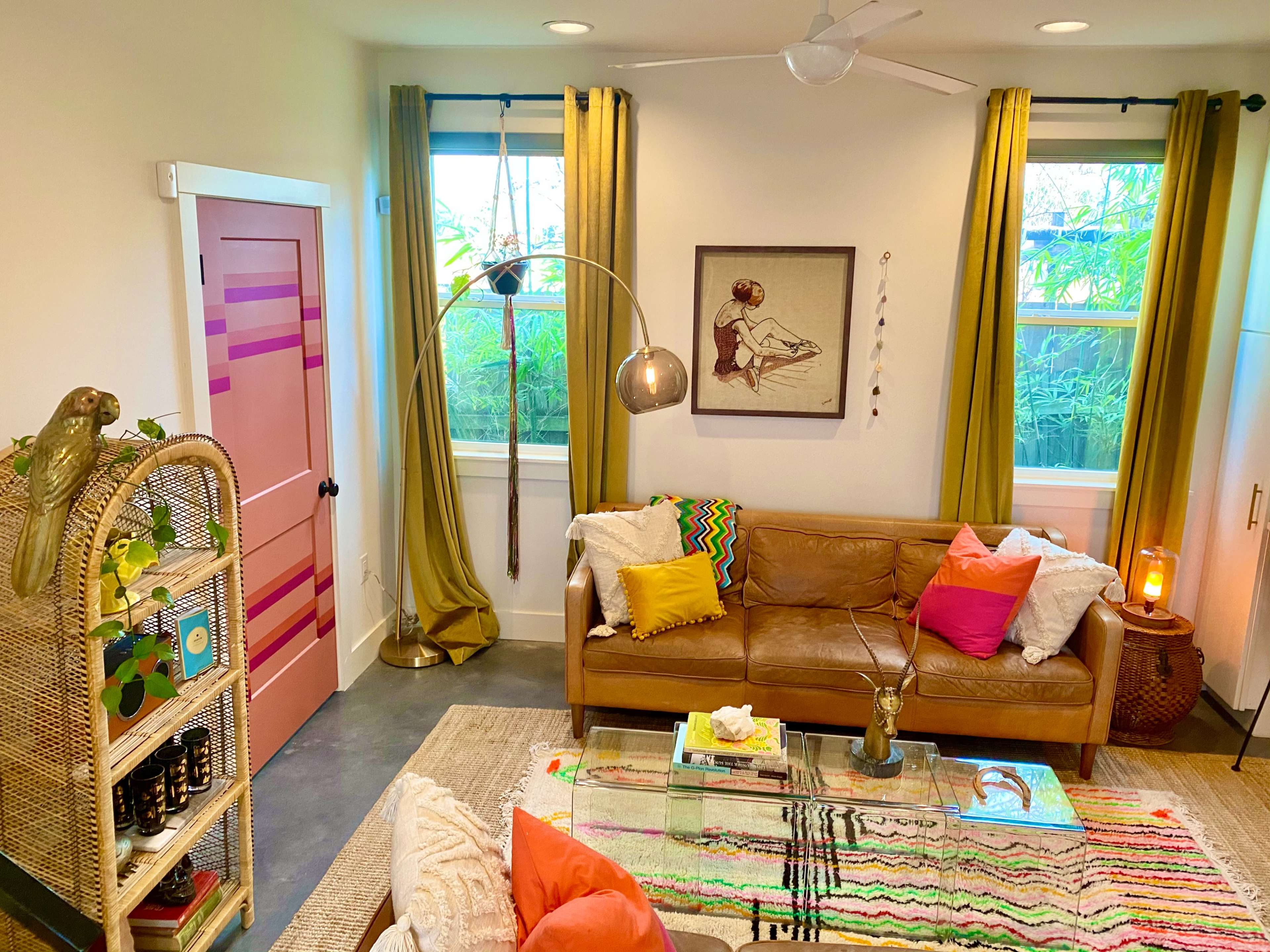The room features a brown leather sofa with colorful throw pillows, a glass coffee table, and large windows with yellow curtains, alongside a pink door and decorative elements.
