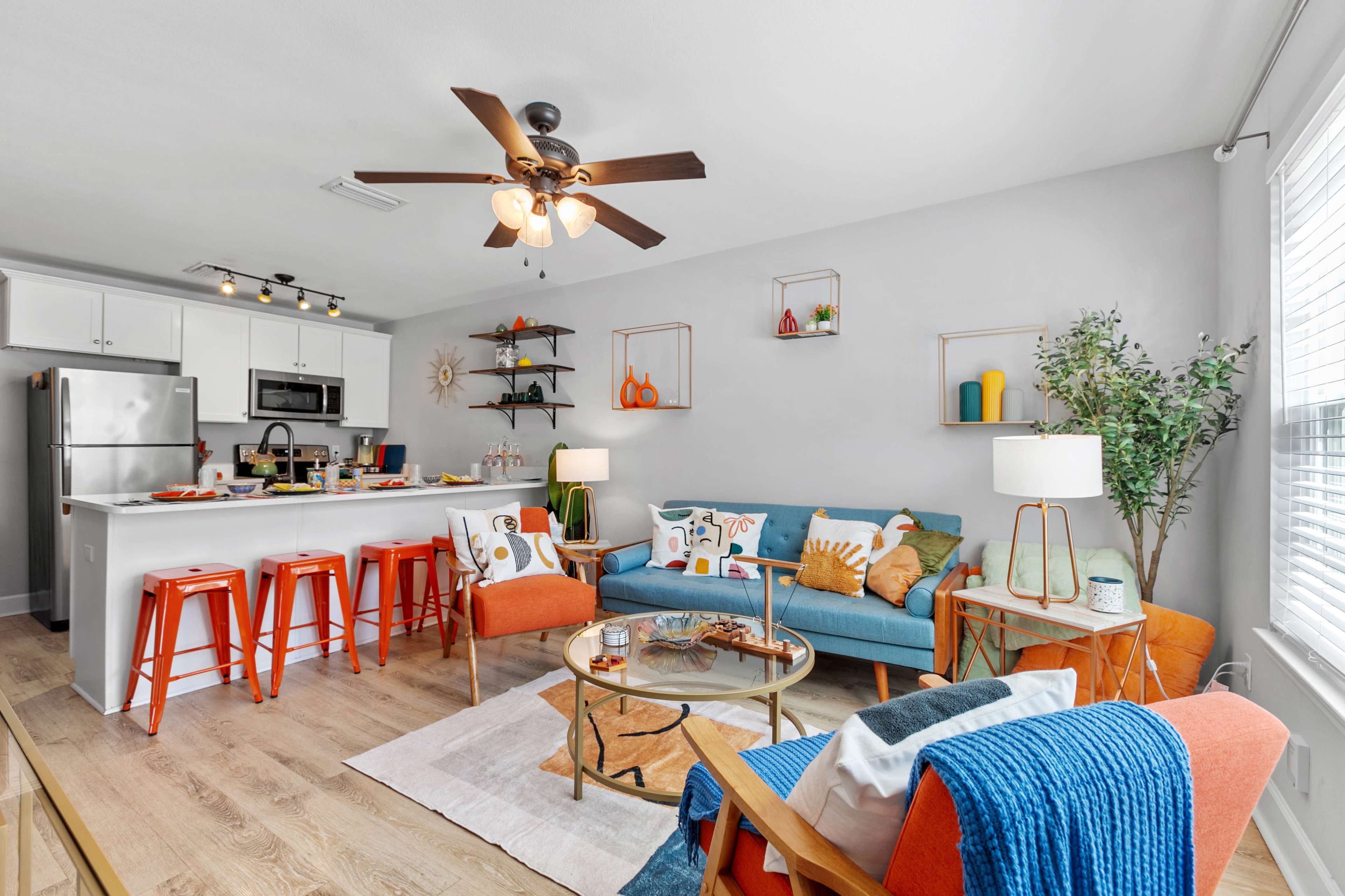A modern living room and kitchen area with a blue sofa, orange bar stools, and various decorative items on shelves and tables.