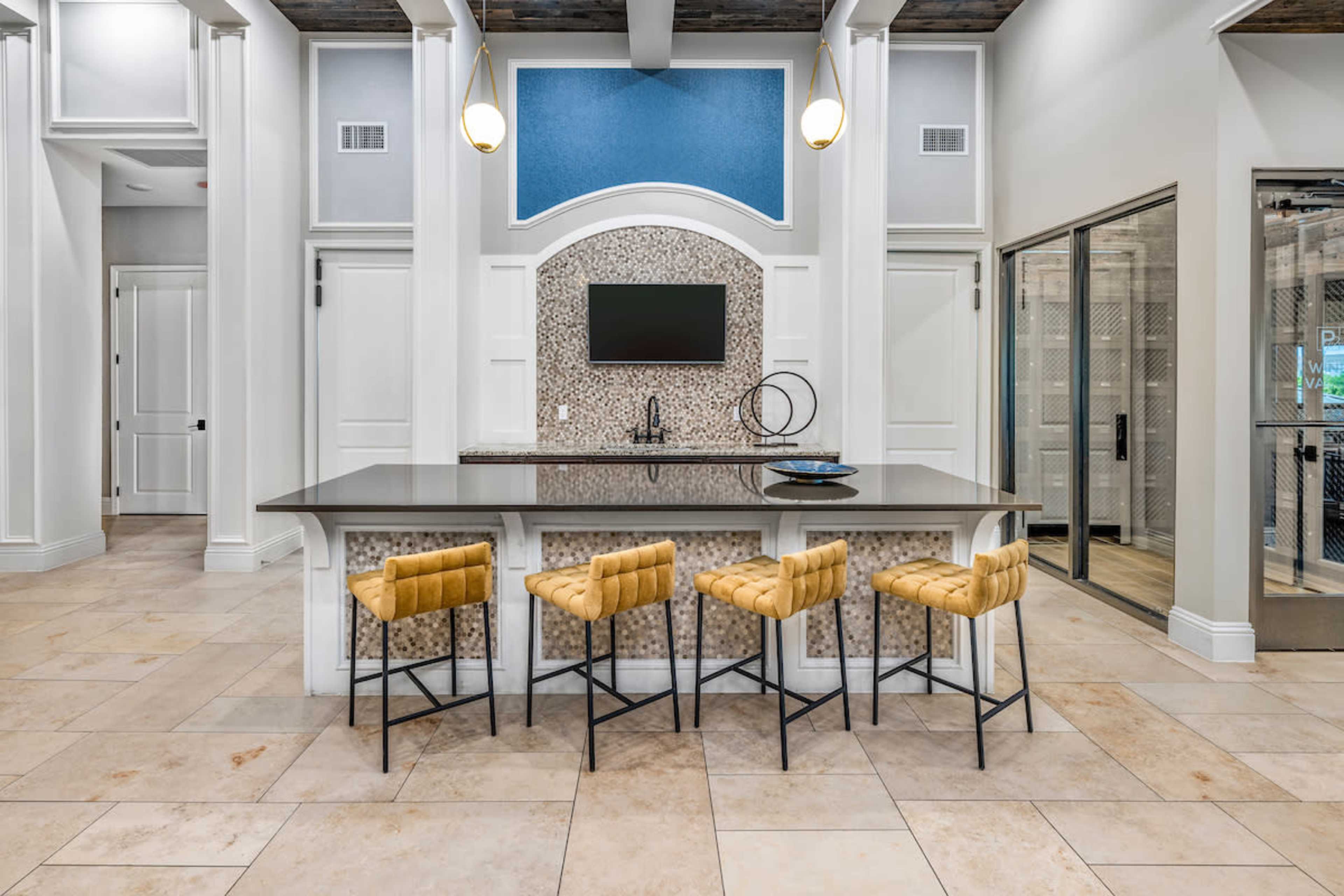 The image shows a modern kitchen island with four yellow bar stools, a flat-screen television mounted on the wall, and decorative tiles on the front of the island.