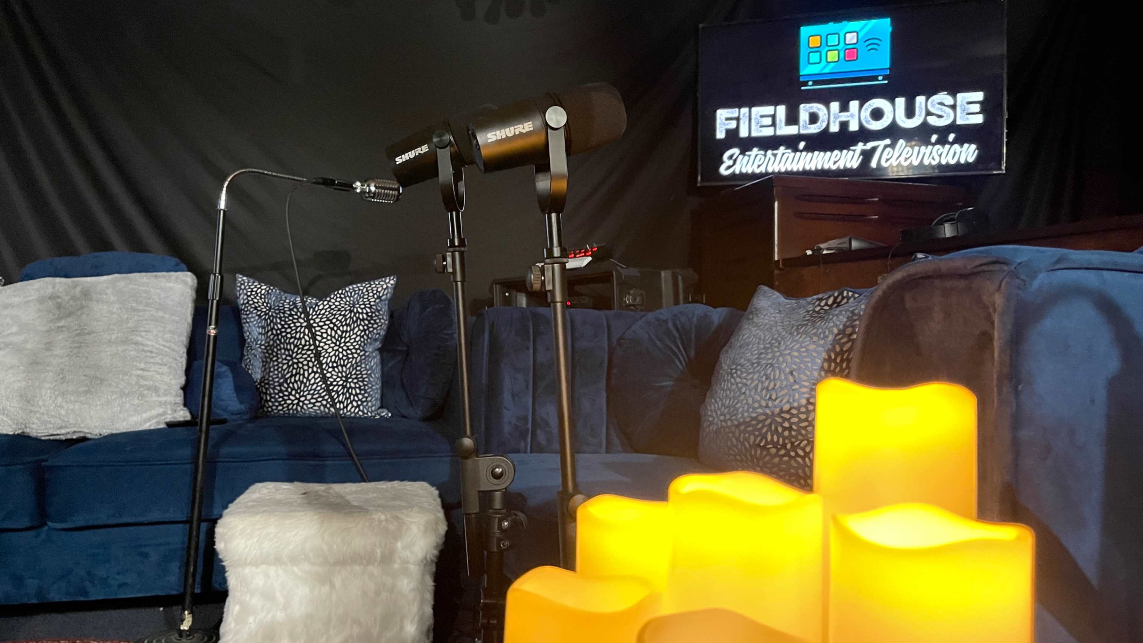 A cozy studio setup features blue couches, two microphones on stands, and LED candles glowing in the foreground.