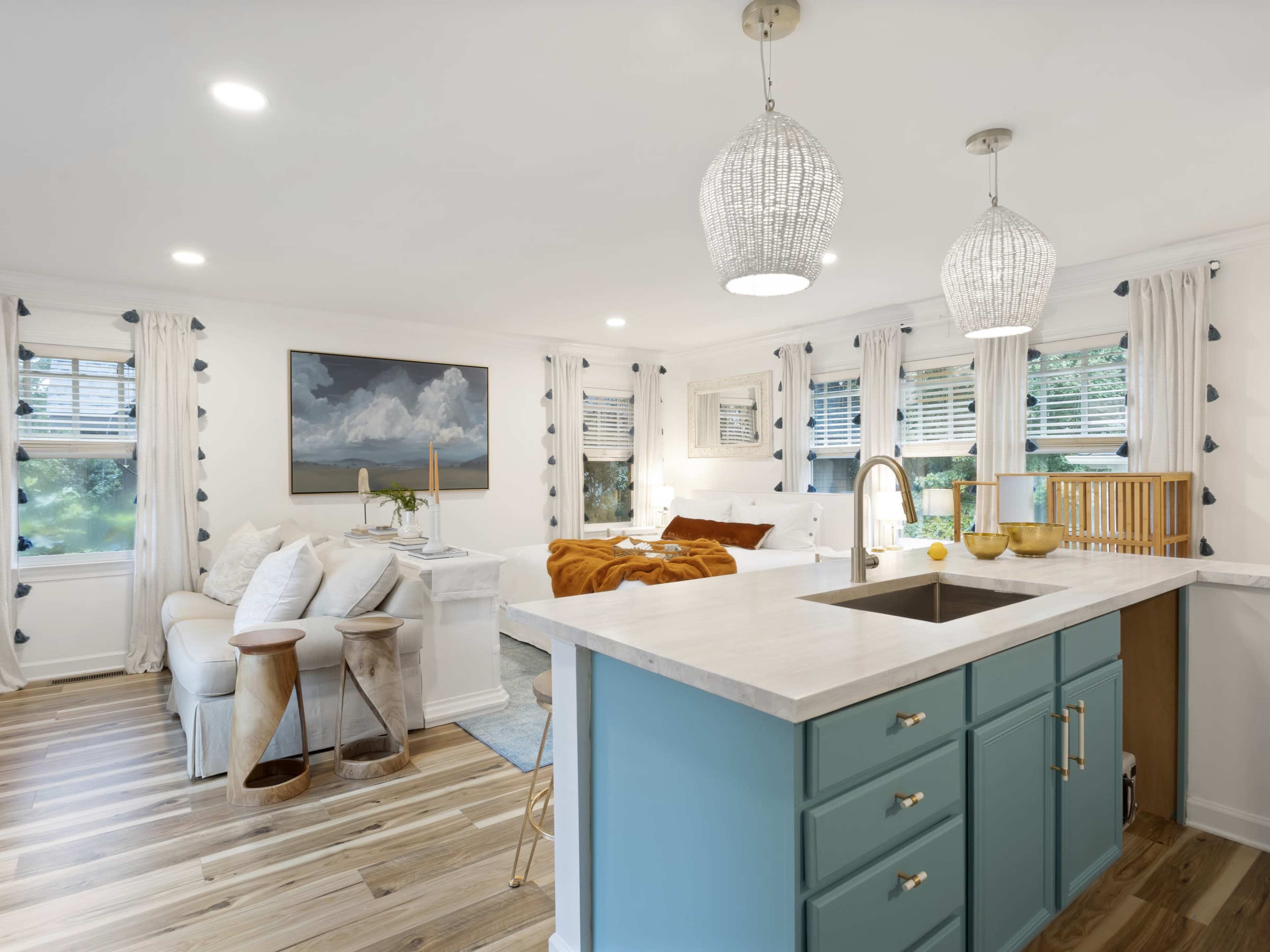 A modern kitchen and living area featuring a central island with a marble countertop, teal cabinetry, and bright natural light streaming through multiple windows.