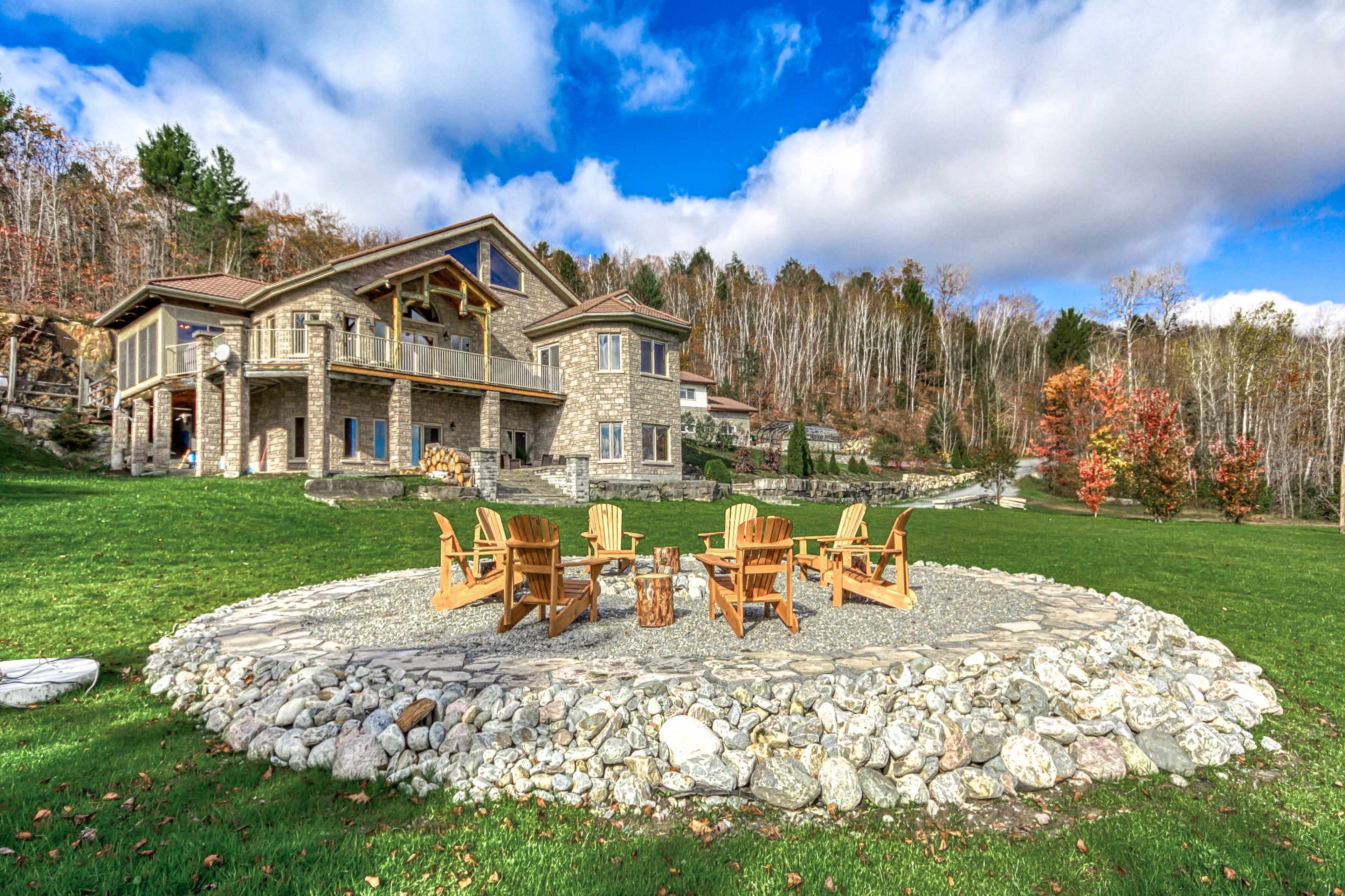 A stone patio with a circular seating arrangement of wooden chairs is situated in front of a large stone house, surrounded by a lush green lawn and trees.