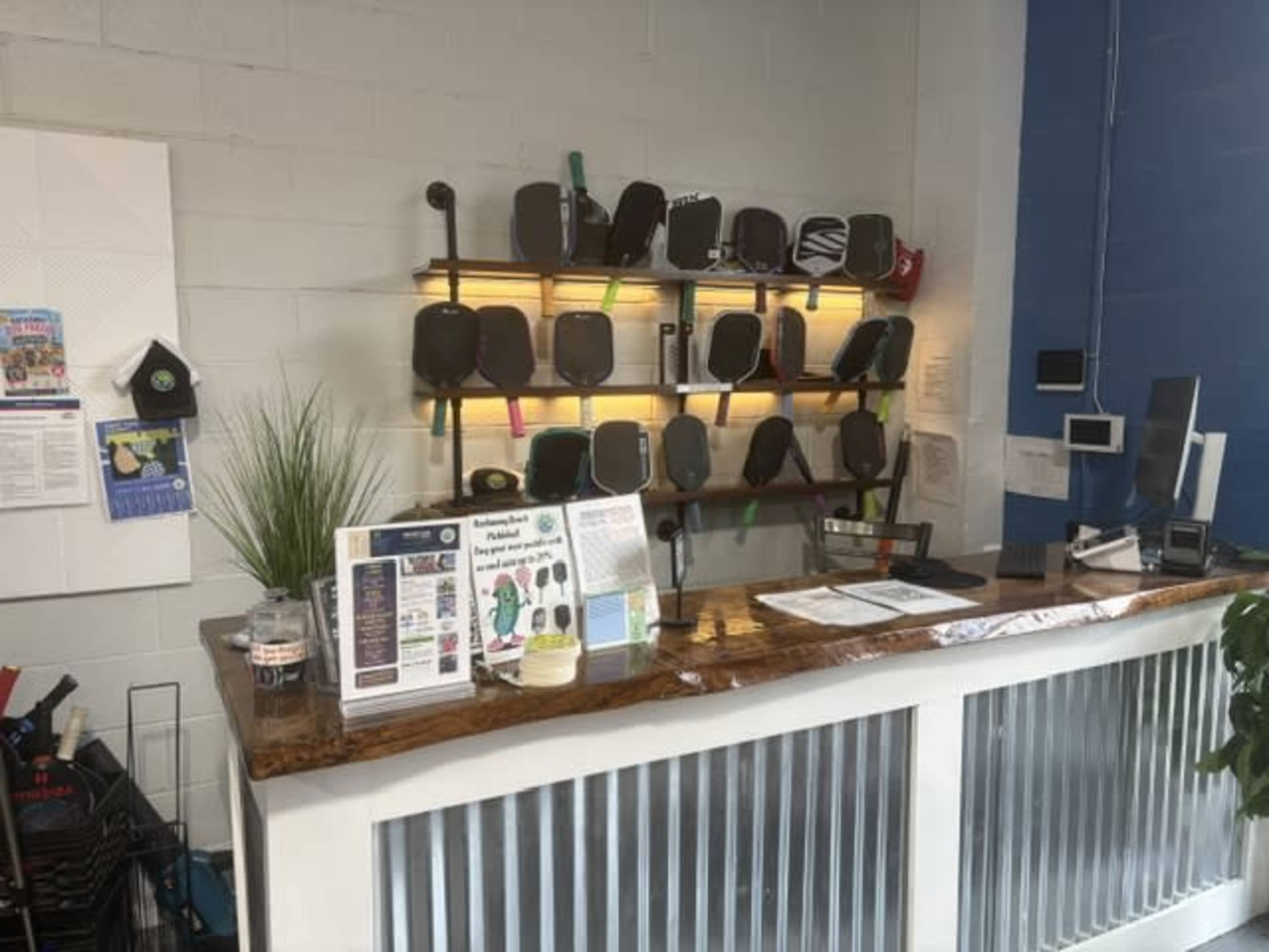 The image shows a reception area with a wooden counter, a display of paddles on a shelf, informational flyers, and a small plant.